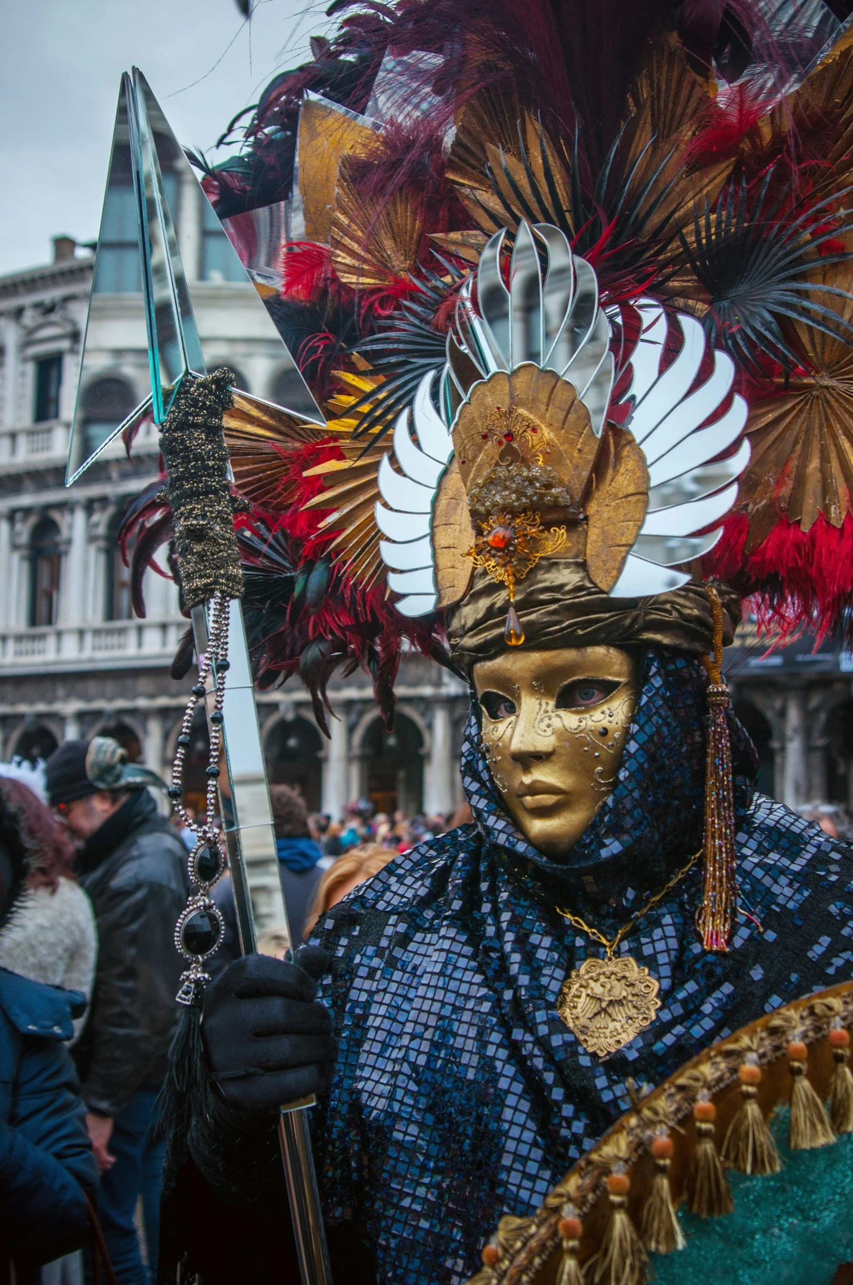 Person in a gold mask and elaborate feathered headdress participating in a parade or festive event.