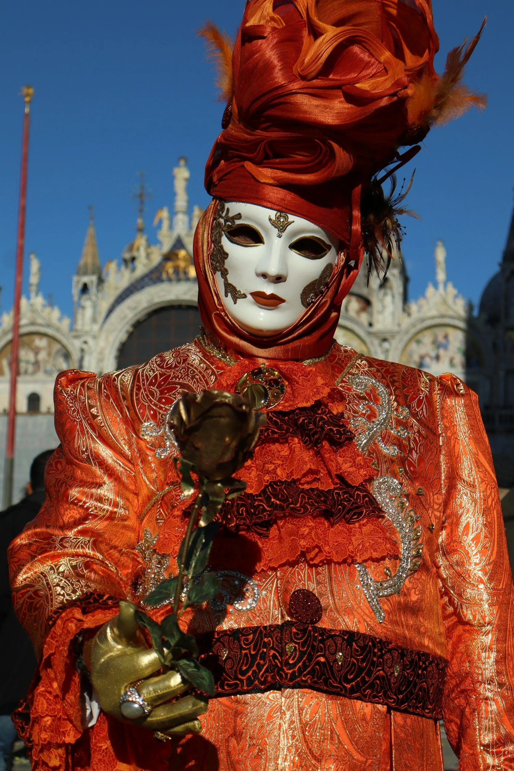 Person dressed in elaborate red and gold costume with a white mask, holding a rose, at Venice Carnival in Venice, Italy.
