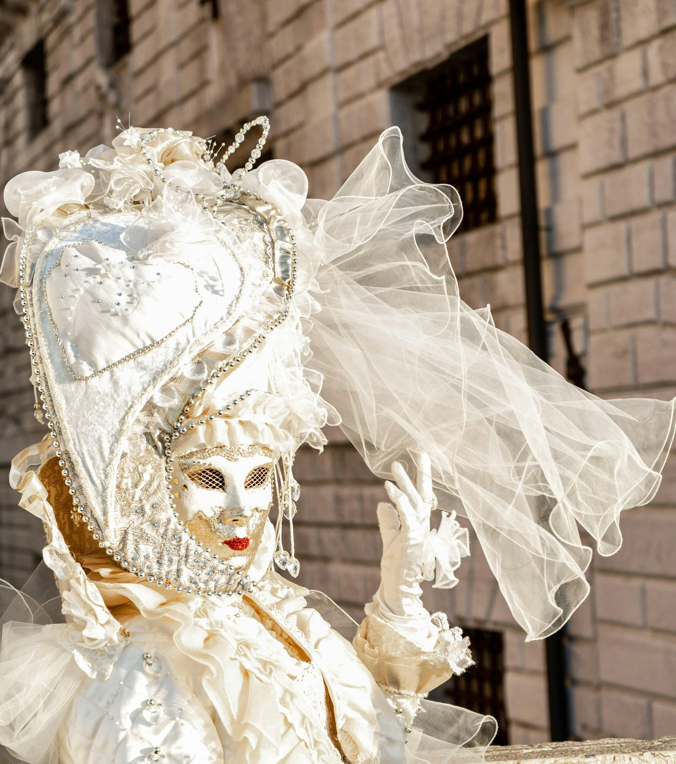 A person in an elaborate white costume and mask, wearing a large, decorated headpiece, standing outdoors with a brick building in the background.