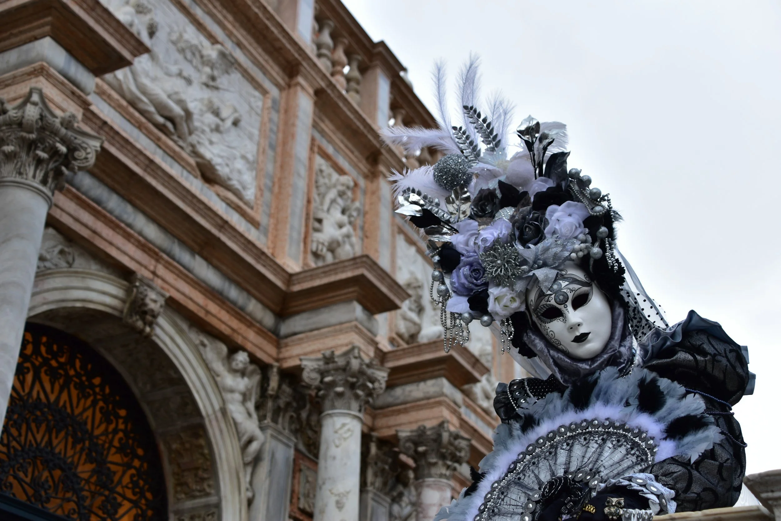 A person dressed in a black and white costume with a mask and elaborate feathered headdress decorated with flowers and ornaments, standing in front of an ornate historic building with detailed stone carvings and columns.