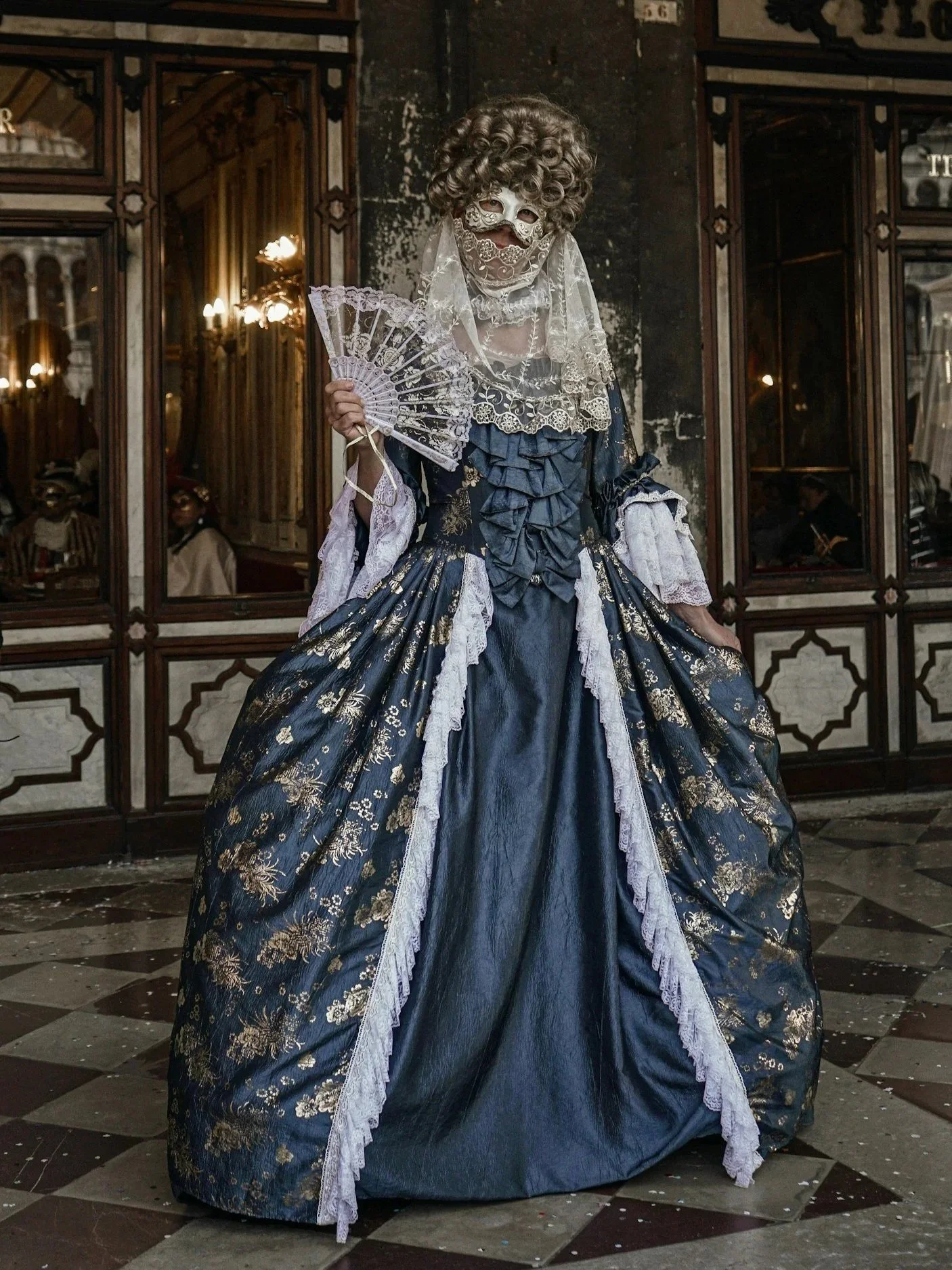 A person wearing an elaborate 18th-century costume with a powdered wig, lace mask, lace veil, blue and gold gown, holding a lace fan, standing indoors.