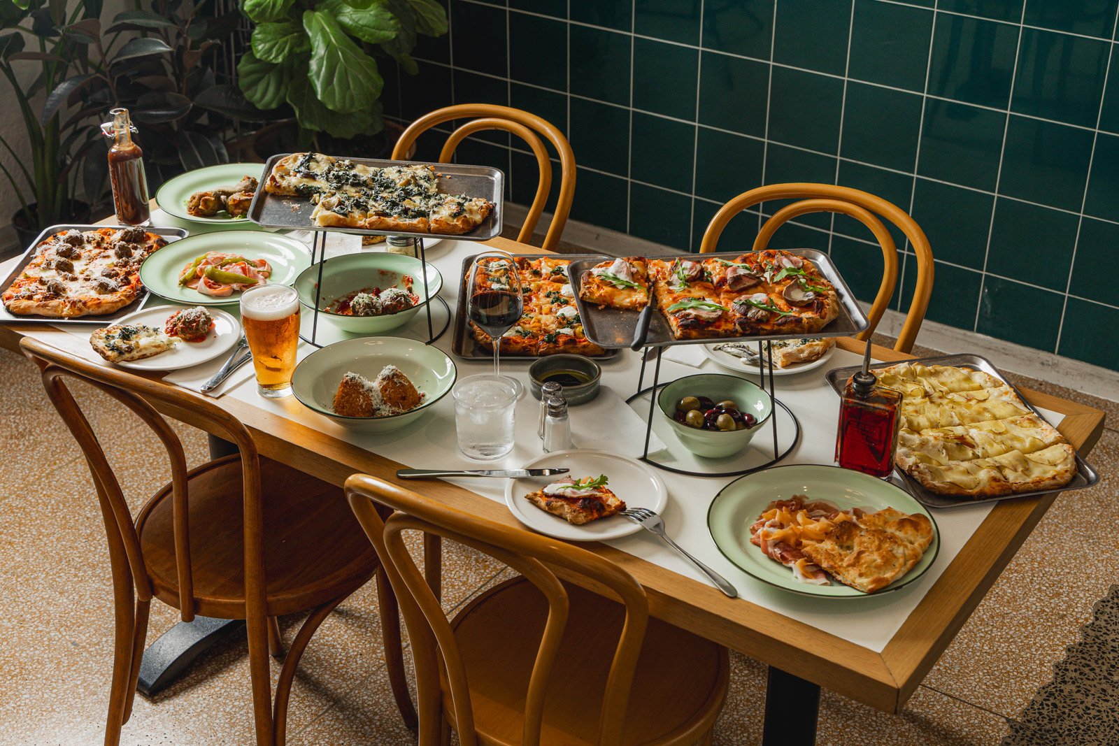 A wooden table set with various pizzas and side dishes, surrounded by wooden chairs, in a restaurant with green tiled walls.