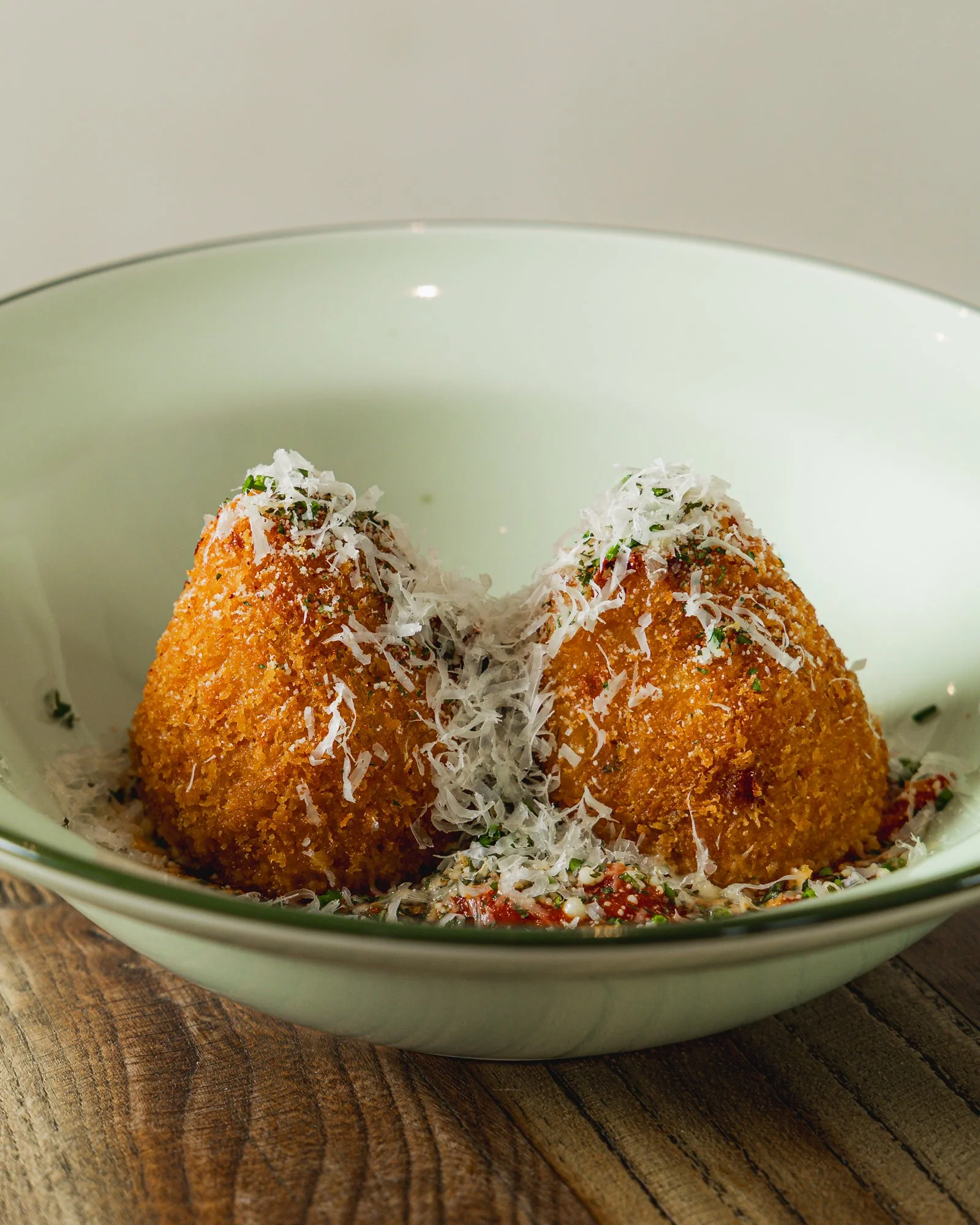 Two breaded and fried arancini served in a bowl, topped with grated cheese and herbs, with a tomato-based sauce underneath.