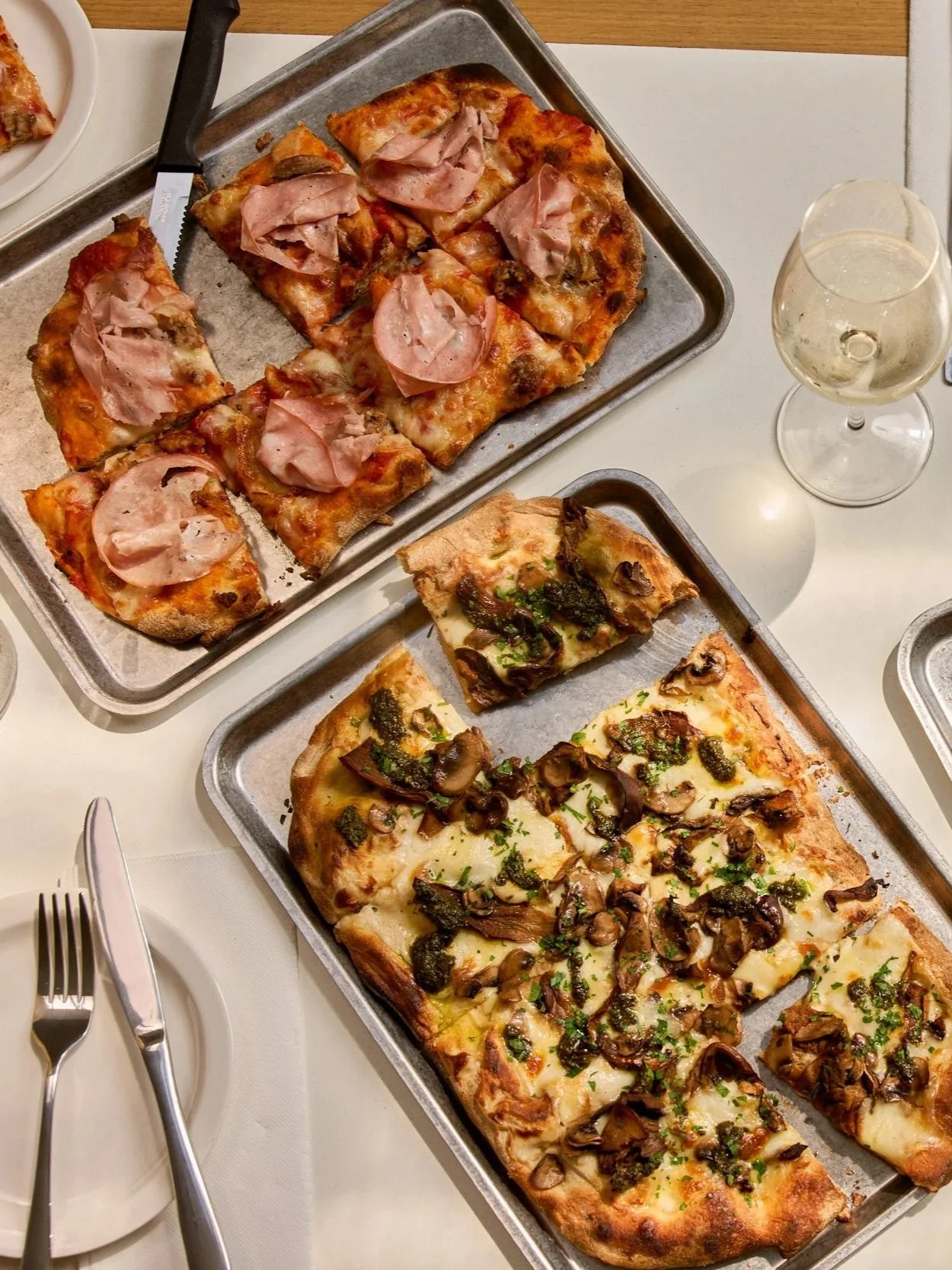 Two trays of freshly baked pizza slices with mushroom and herb toppings, and another with ham and cheese, on a white table with wine glasses.