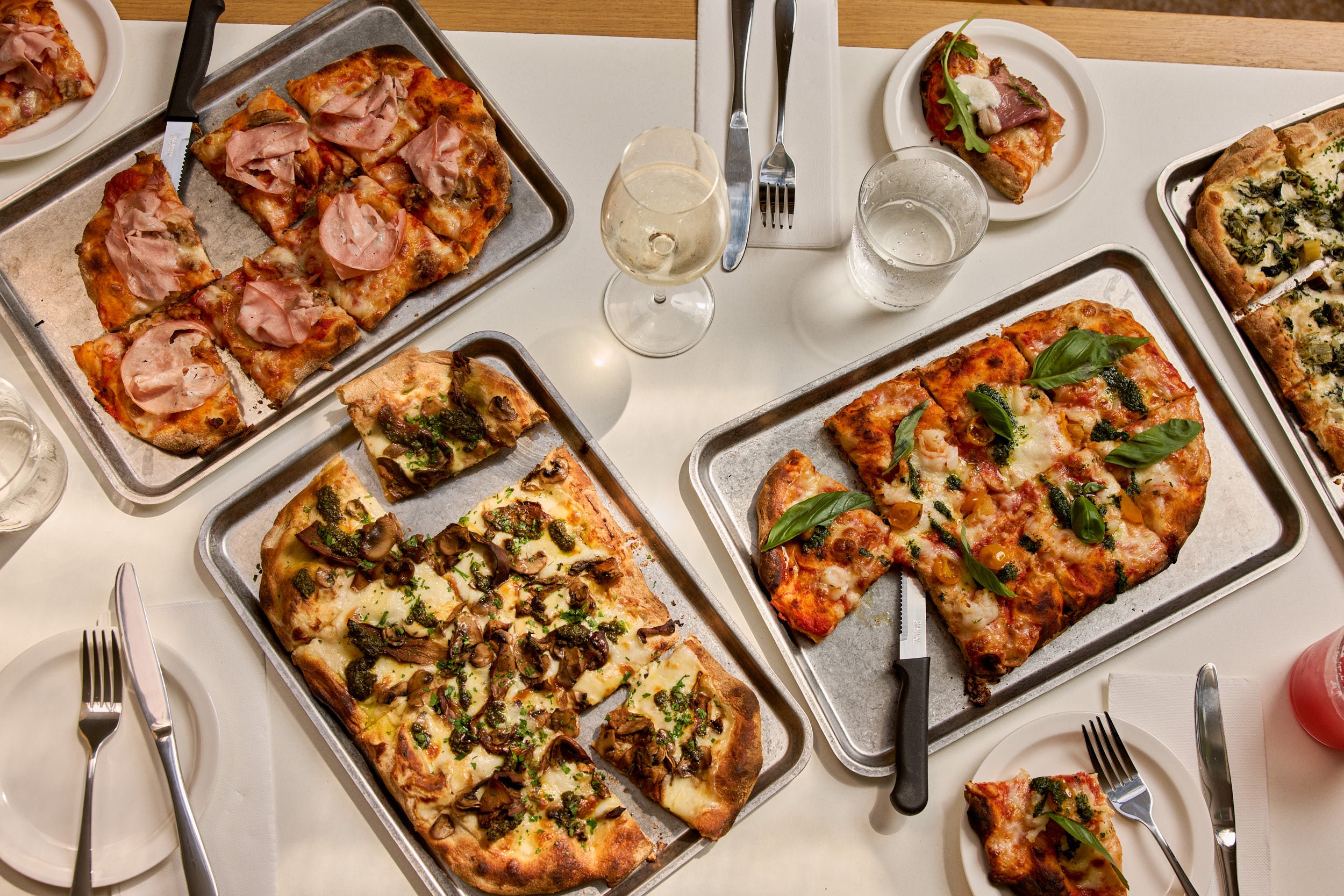Assorted pizzas with various toppings on metal trays, set on a white table with wine glasses, water glasses, and plates.