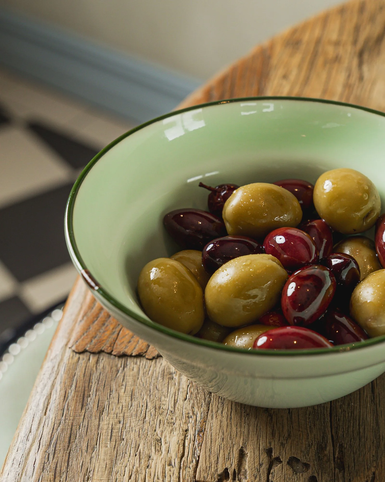 A green and white bowl filled with green and purple olives sitting on a rustic wooden table.