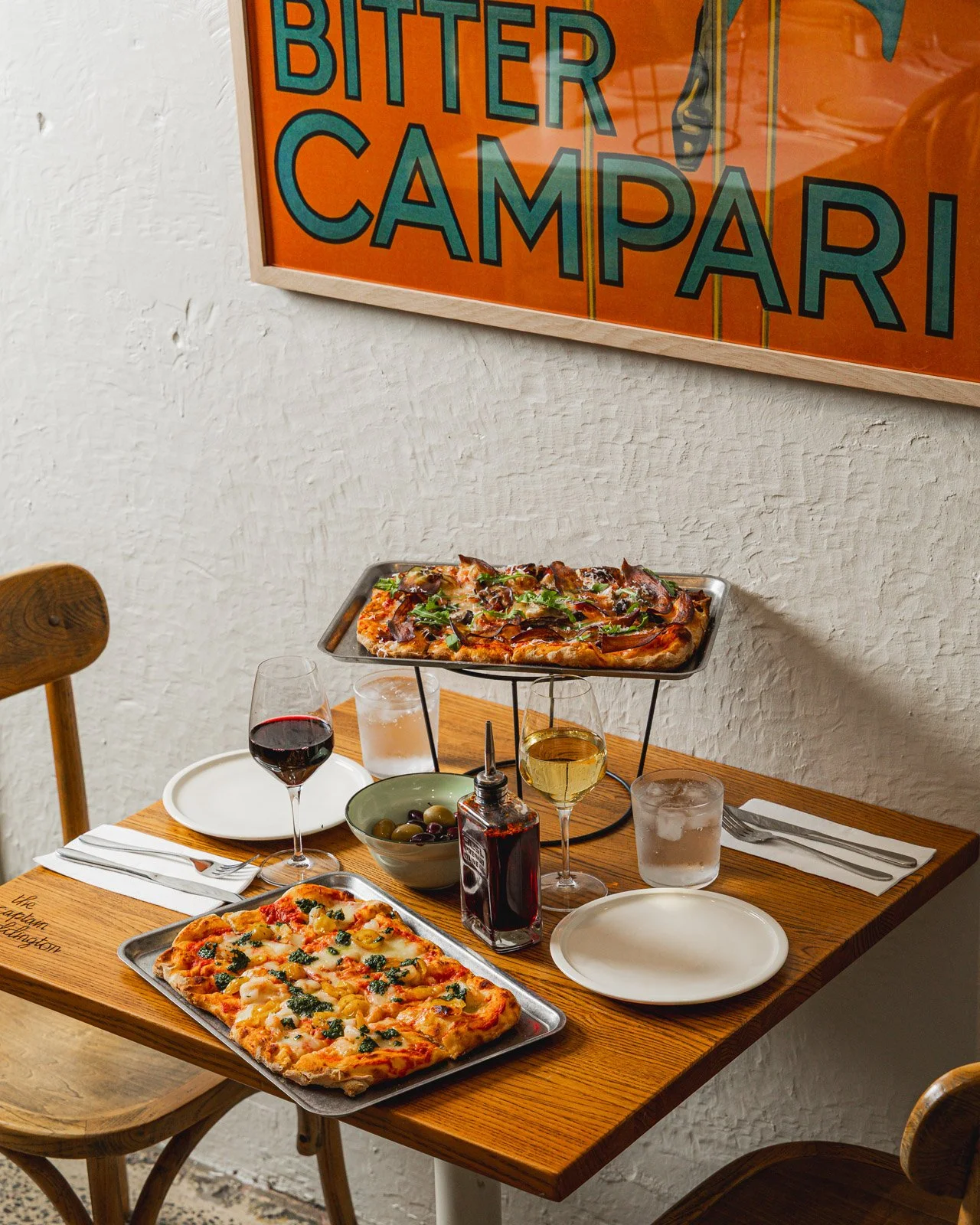A table set for two with two pizzas, a glass of red wine, a white wine glass, a glass of water, a bowl of olives, a bottle of hot sauce, and empty plates and utensils. There is a framed poster on the wall with the words 'Bitter Campari' in colorful letters.