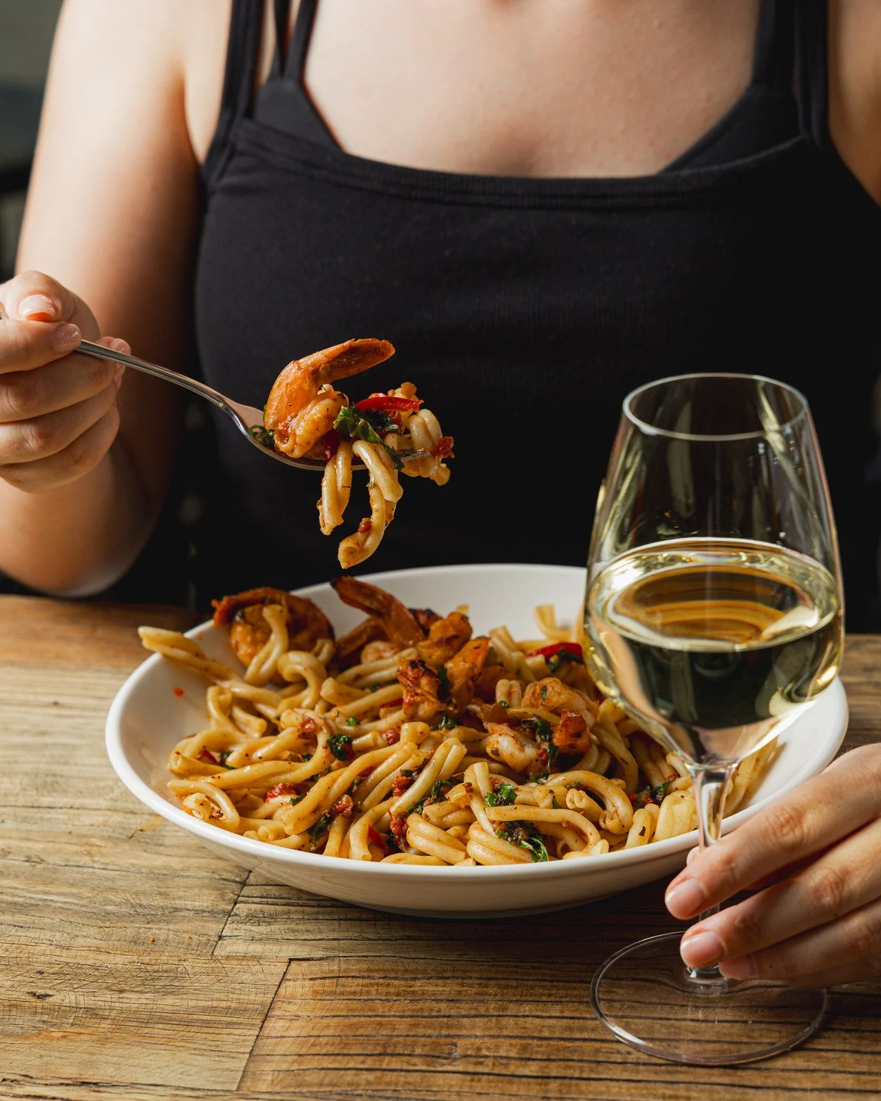 A woman in a black top is eating a plate of pasta with seafood, tomatoes, and herbs, while holding a glass of white wine on a wooden table.