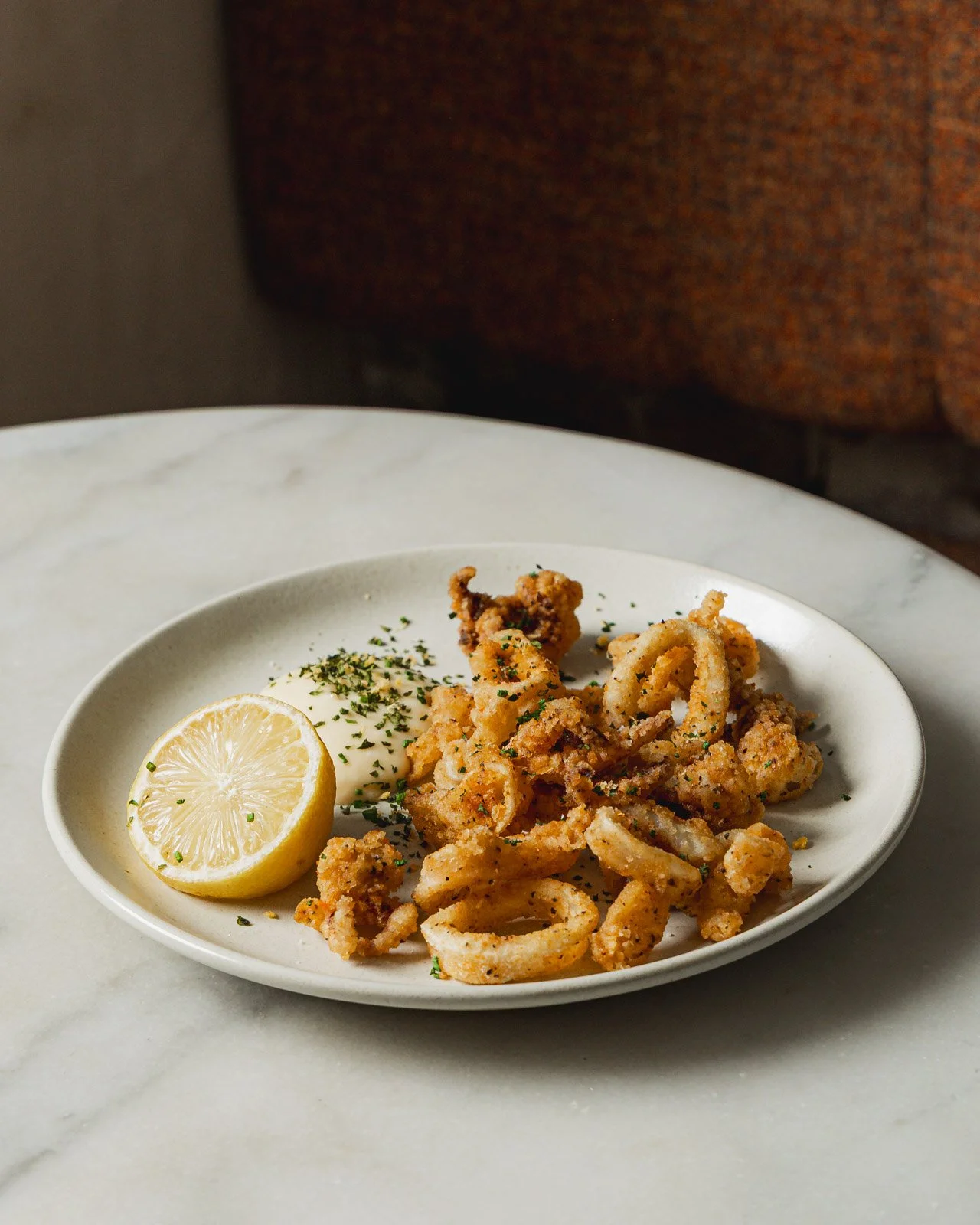 Fried calamari served on a white oval plate with lemon wedge, mayonnaise, and chopped herbs on top, on a marble table.