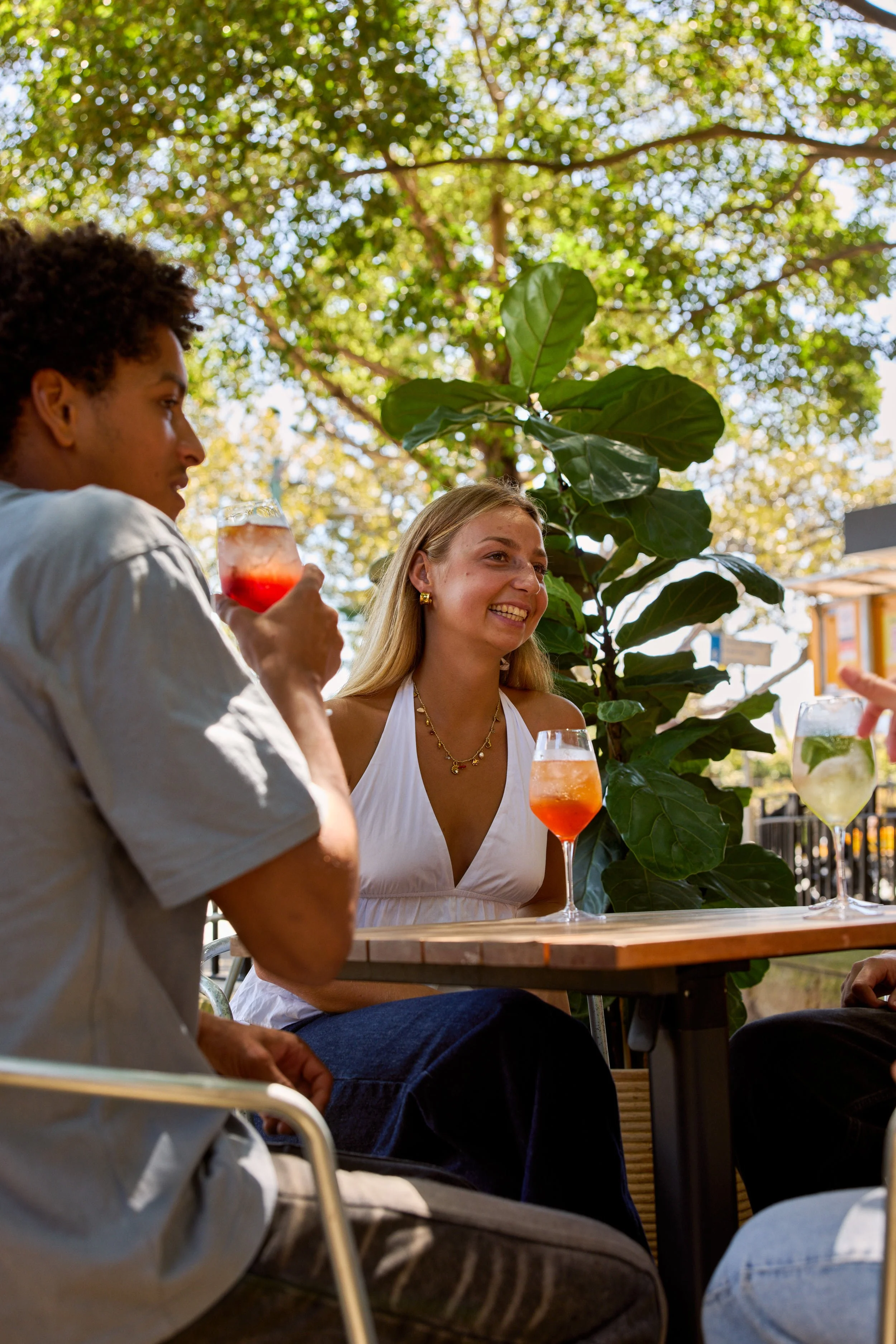 Three friends enjoying drinks at an outdoor cafe table surrounded by greenery and trees, with sunlight filtering through the leaves.