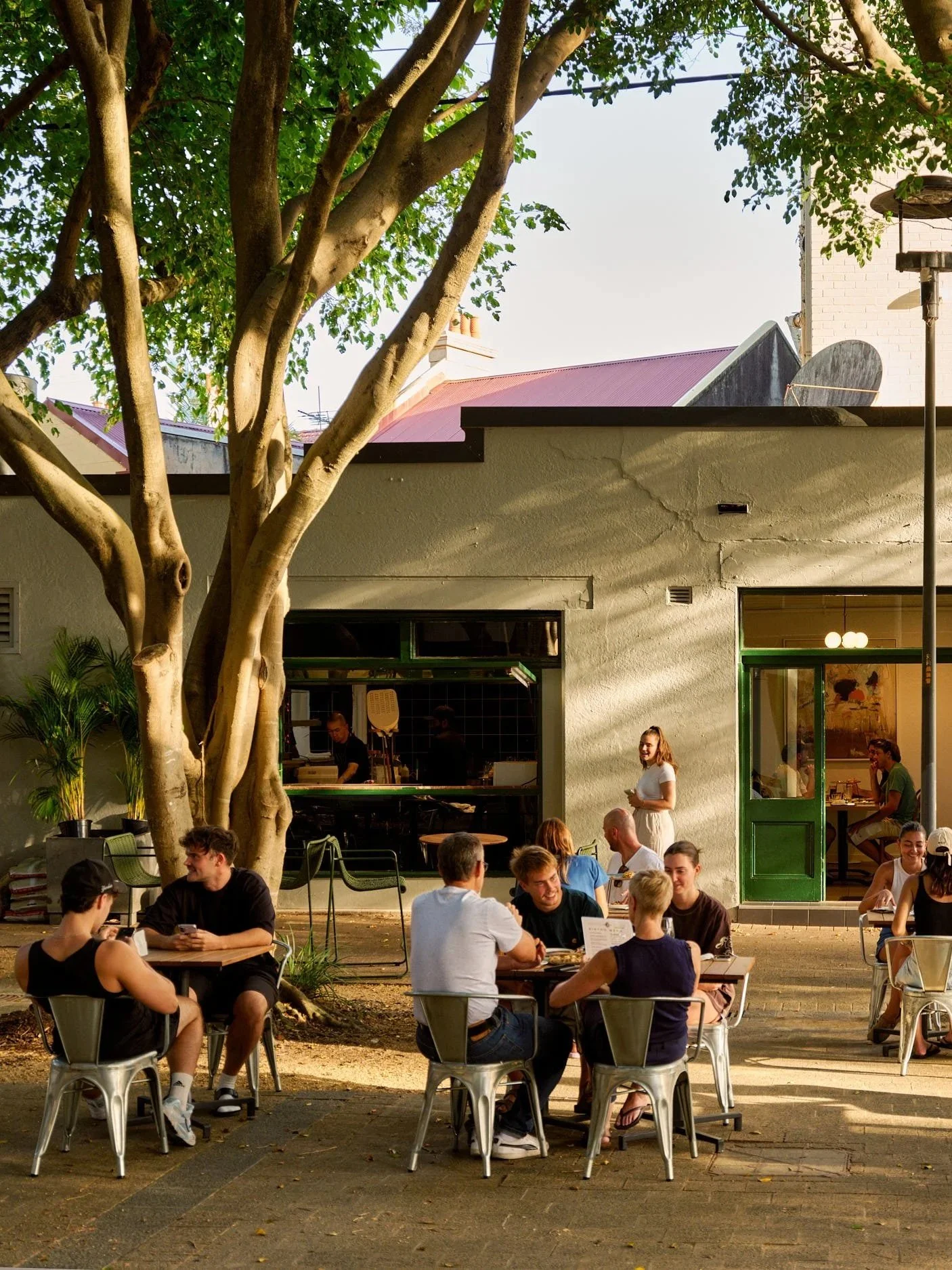 People dining outdoors at a restaurant with a large tree in the foreground, a building with a green door, and a window, in the evening or late afternoon.