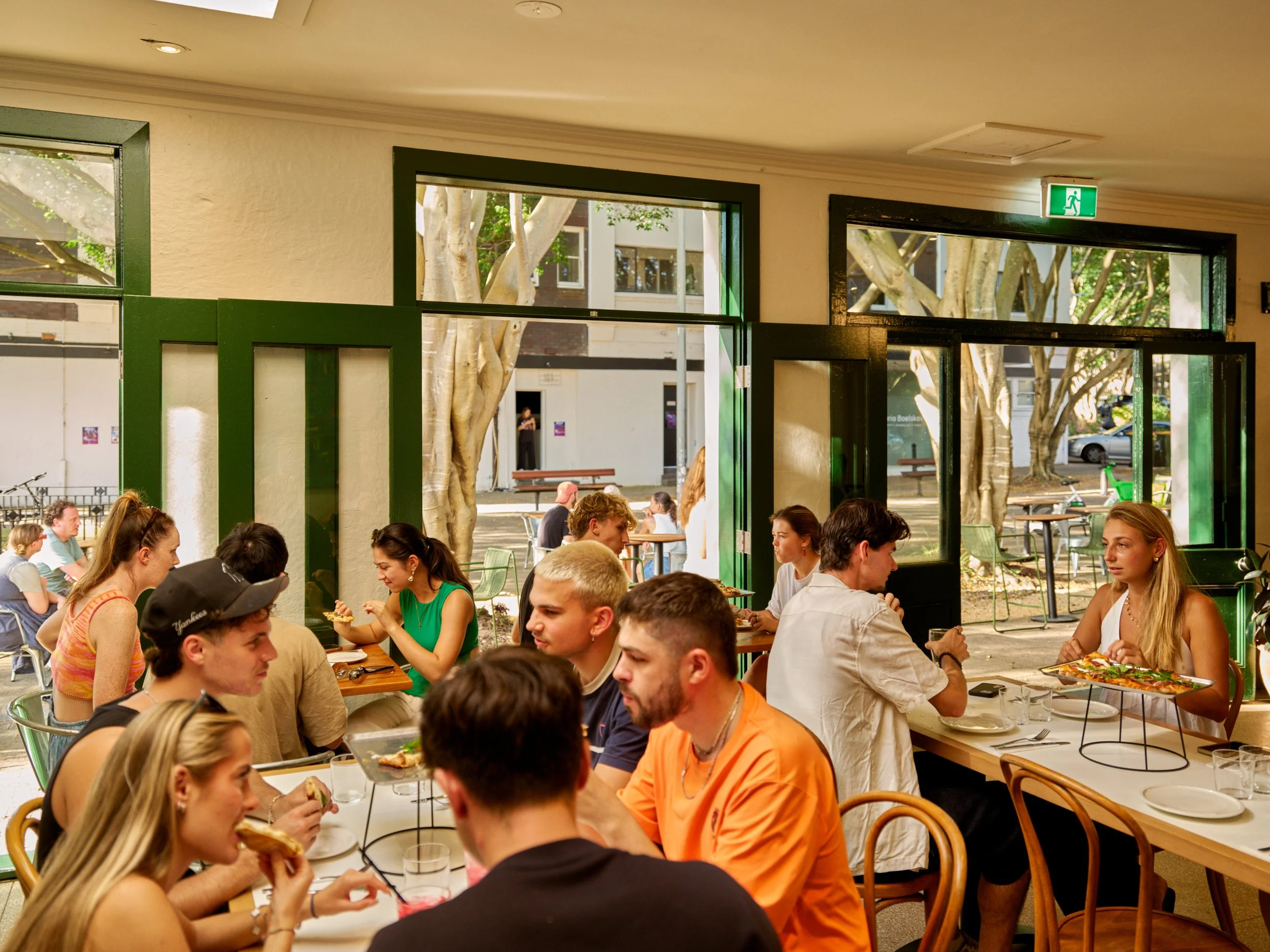 People dining inside a restaurant with large windows overlooking outdoor seating and trees.