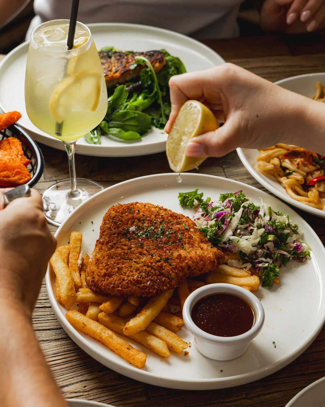 A plate of fried chicken with French fries, coleslaw, and dipping sauce, with a lemon wedge being squeezed over it, surrounded by other dishes and a tropical drink on a wooden table.
