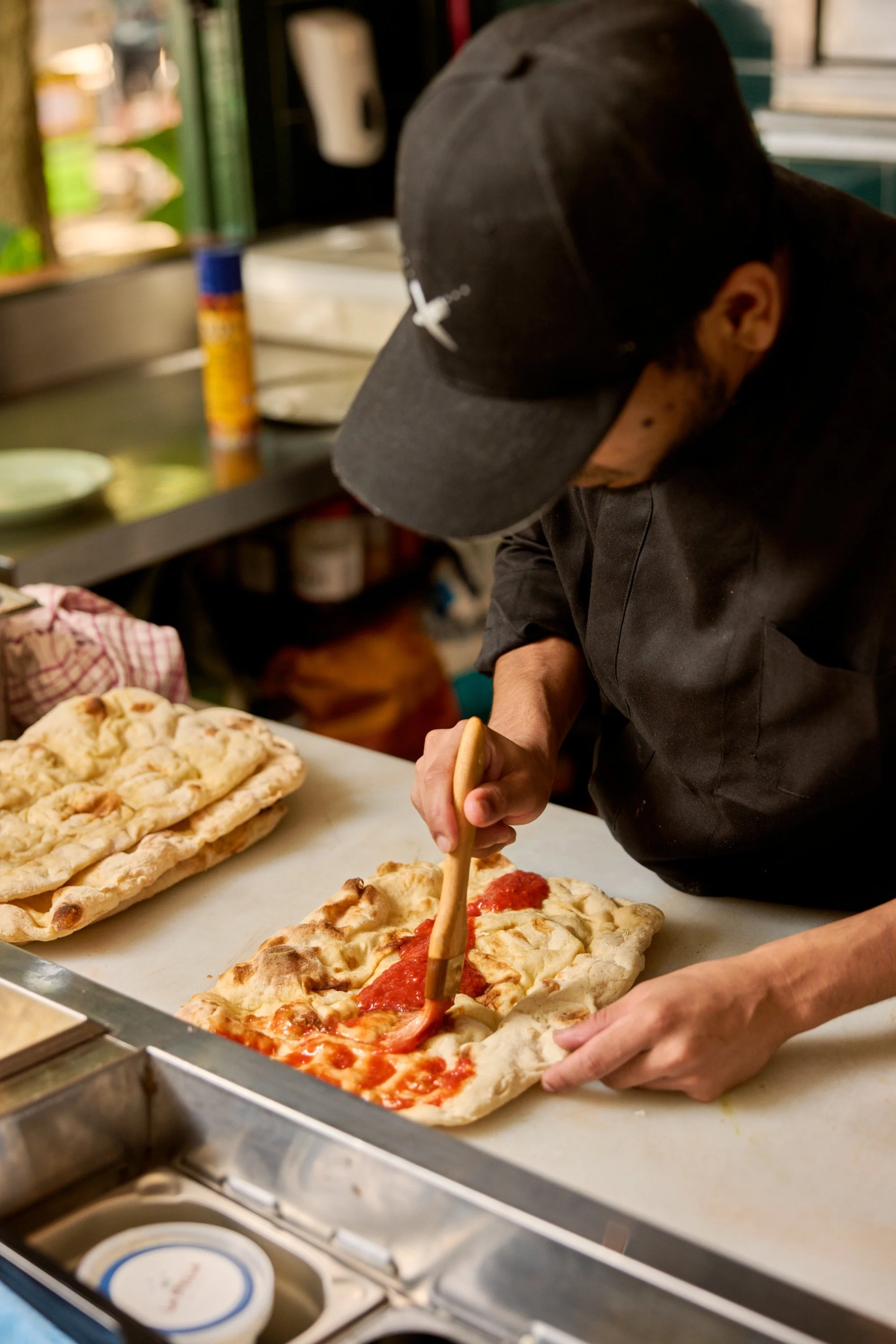 A person wearing a black cap and black shirt making a pizza by spreading sauce on a rectangular pizza crust at a kitchen counter.