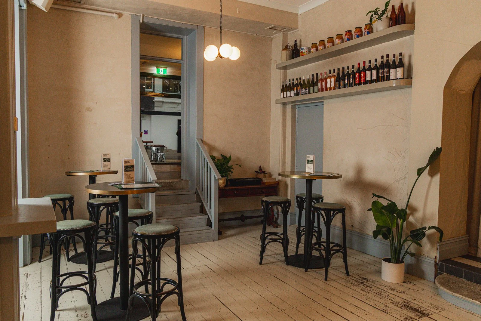 Interior of a cozy paddington pub with high tables, bar stools, a staircase, a shelf with wine bottles, potted plants, and warm lighting.
