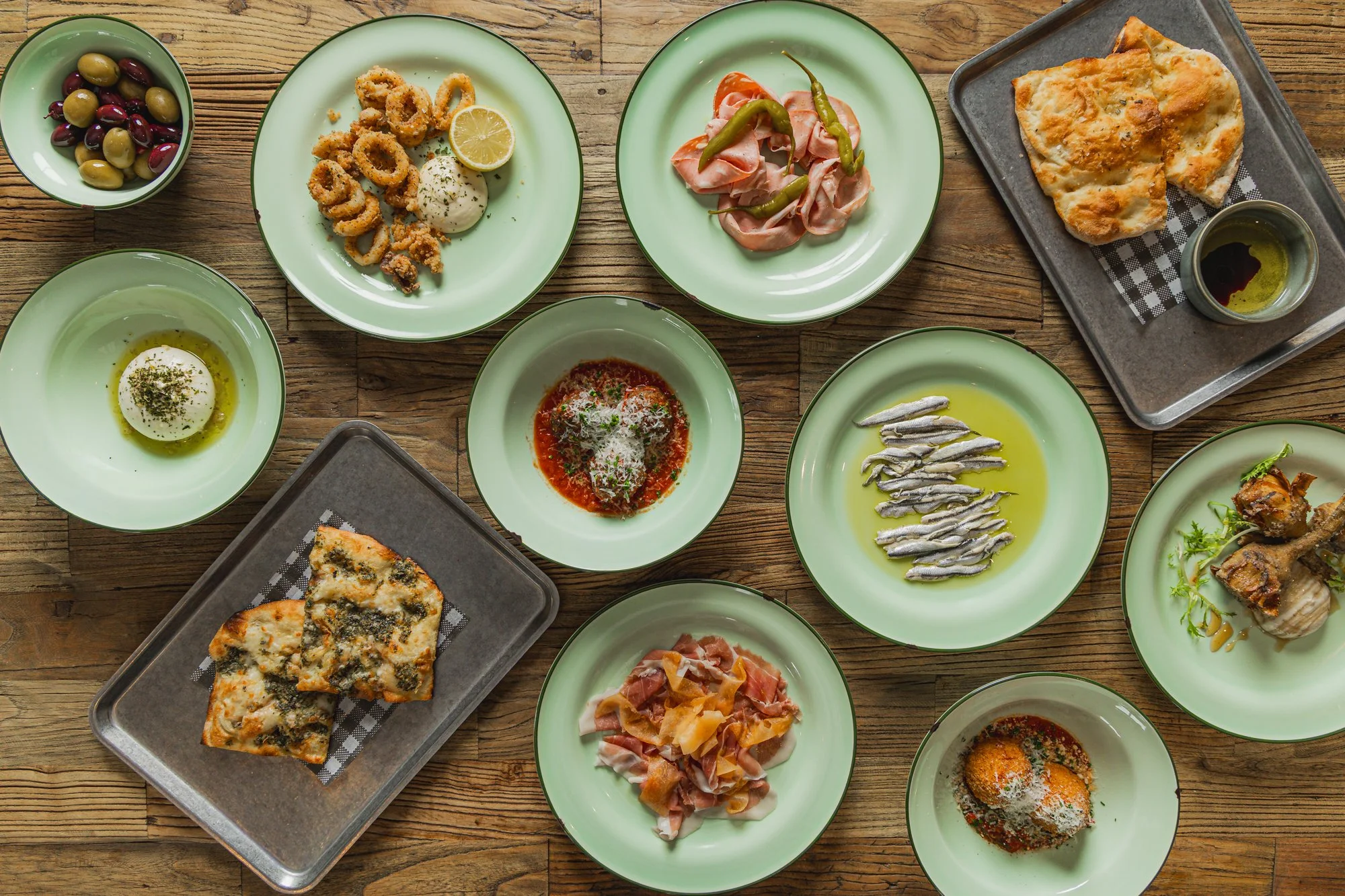 A variety of Mediterranean dishes on green and gray plates and bowls on a wooden table, including olives, calamari, prosciutto, pizza, anchovies, fried eggs, meatballs, and bread with dip.