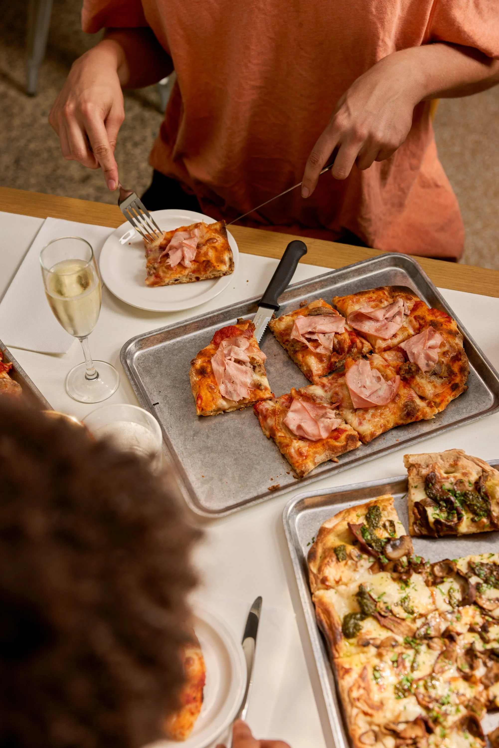 Person in orange shirt cutting a slice of pizza topped with ham from a metal tray, with other pizza slices on the table, along with a glass of white wine.