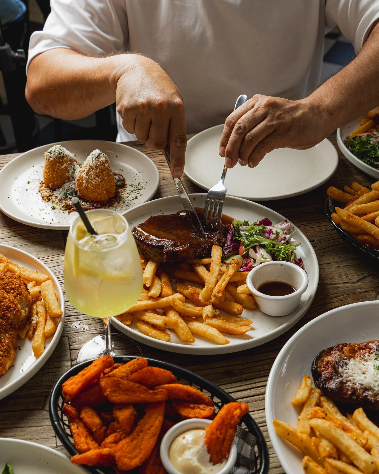 People eating a meal featuring steak, fries, salad, fried appetizers, and drinks at a wooden table.