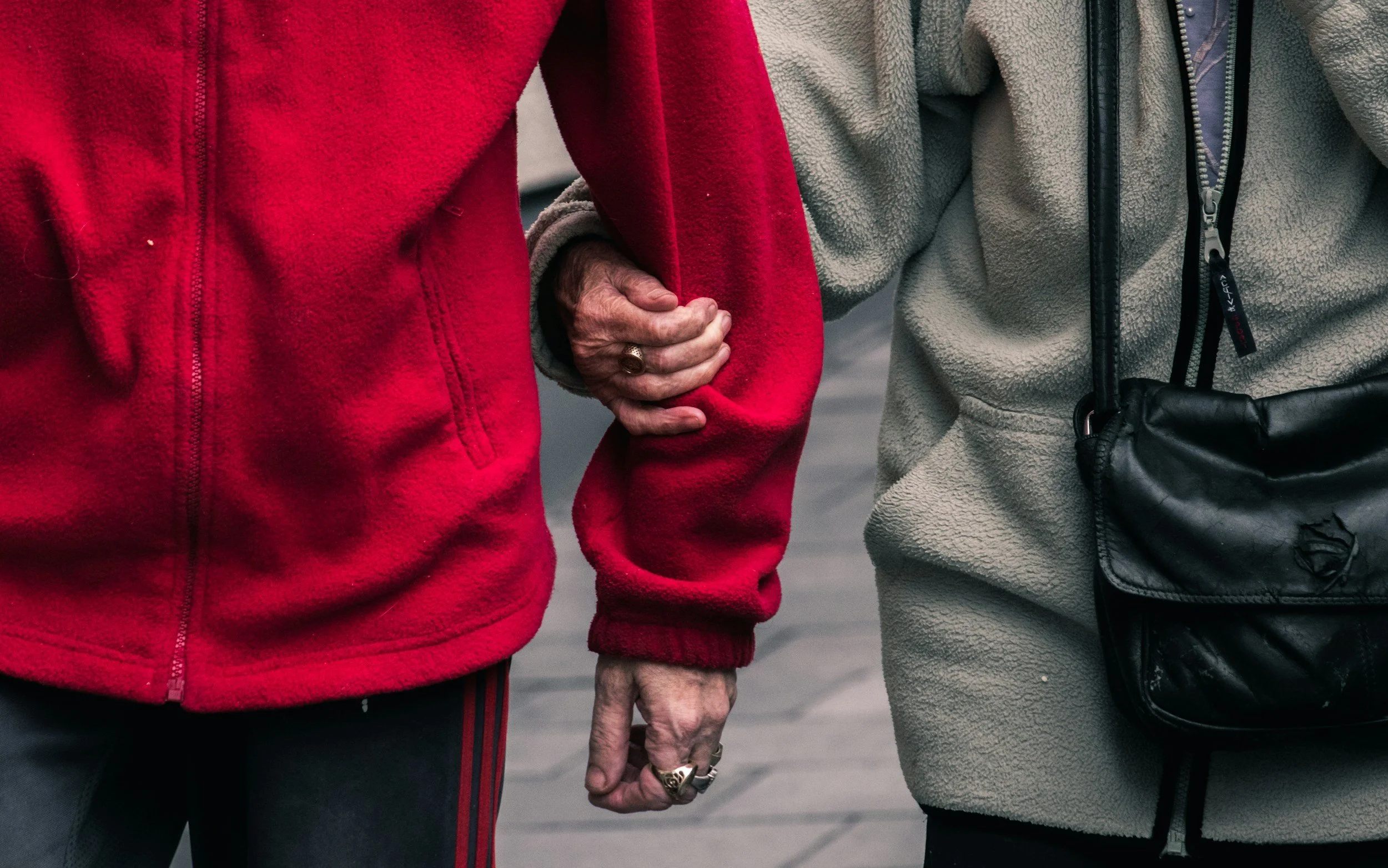 Two people holding hands, one wearing a red fleece jacket and the other wearing a beige fleece jacket with a black bag.