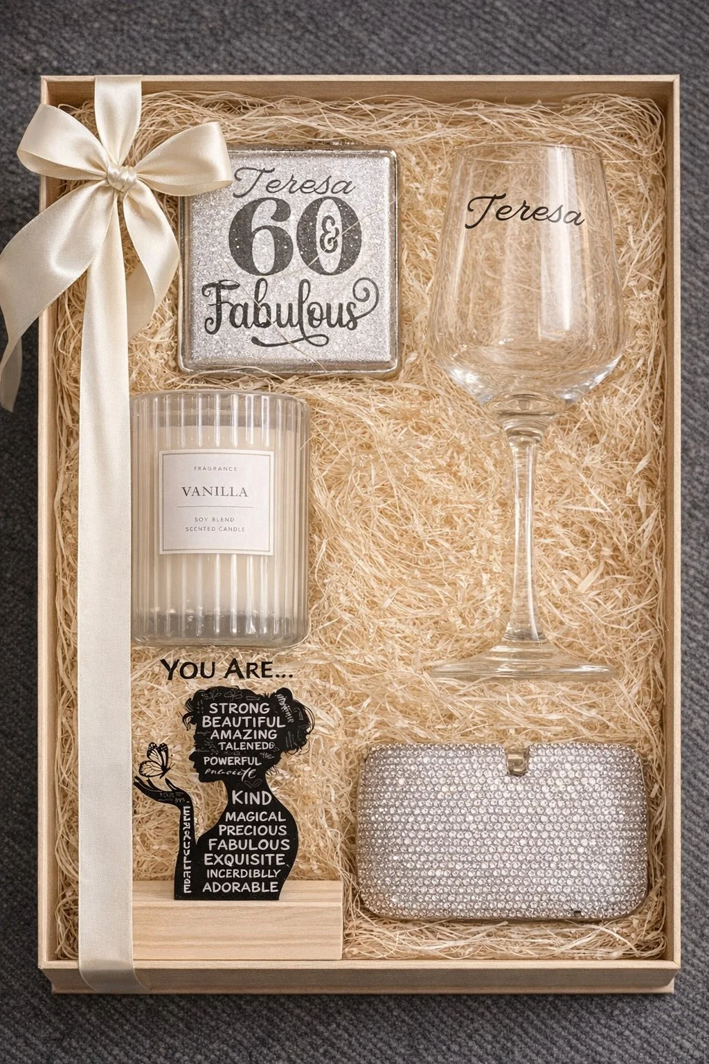 Gift box with a silver '60 and fabulous' plaque, an empty wine glass engraved with 'Teresa', a vanilla scented candle, a silver clutch, and a decorative silhouette with positive words, all arranged on shredded paper and tied with a cream ribbon.