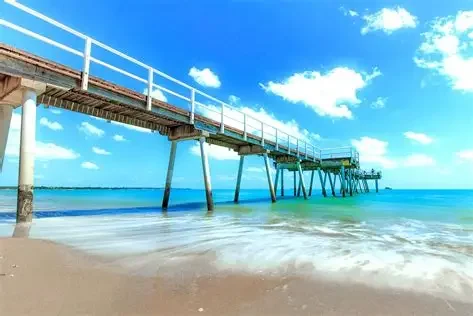 A wooden pier extends over the ocean with a blue sky and white clouds above, and gentle waves hitting the sandy shore below.
