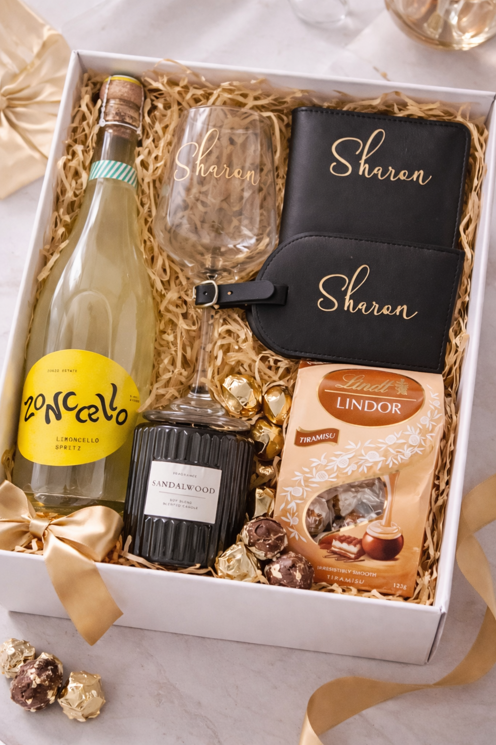 A gift box containing a bottle of lemoncello spritz, a wine glass with the name 'Sharon', a black notebook and wallet with 'Sharon' written on them, a scented candle labeled 'Sandalwood', a box of Lindt Lindor Tiramisu chocolates, and some gold-wrapped candies, all arranged on shredded paper with a gold ribbon.