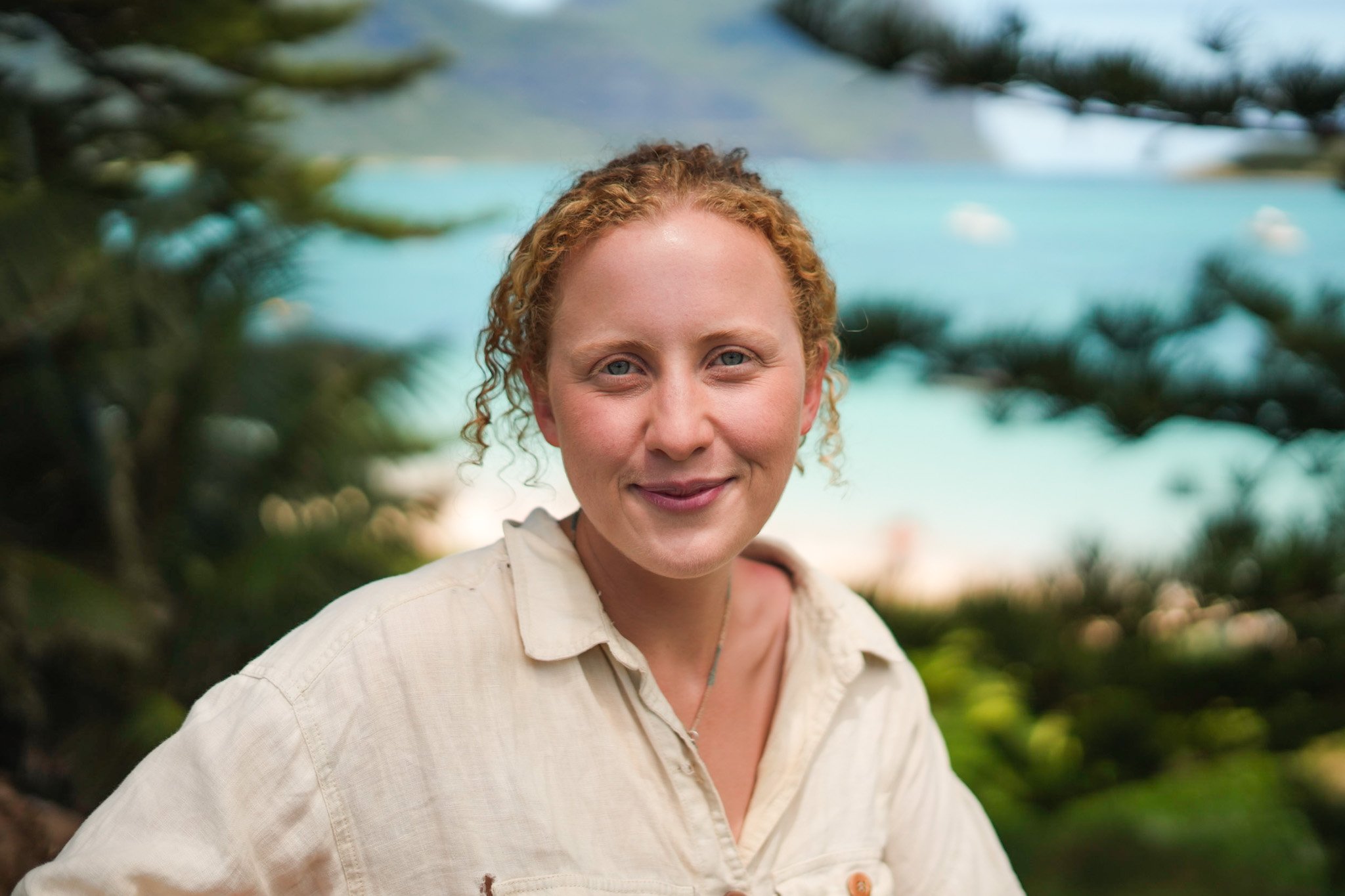 A woman with curly red hair outdoors, smiling, with a background of trees and a body of water.