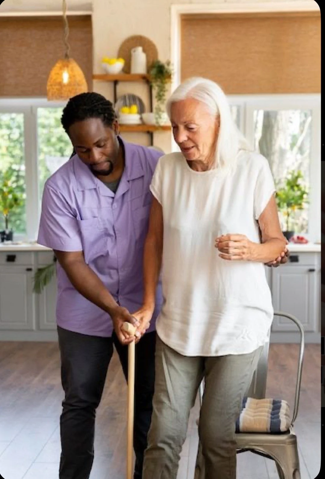 A caregiver assists an elderly woman with walking using a cane in a cozy, well-lit kitchen.