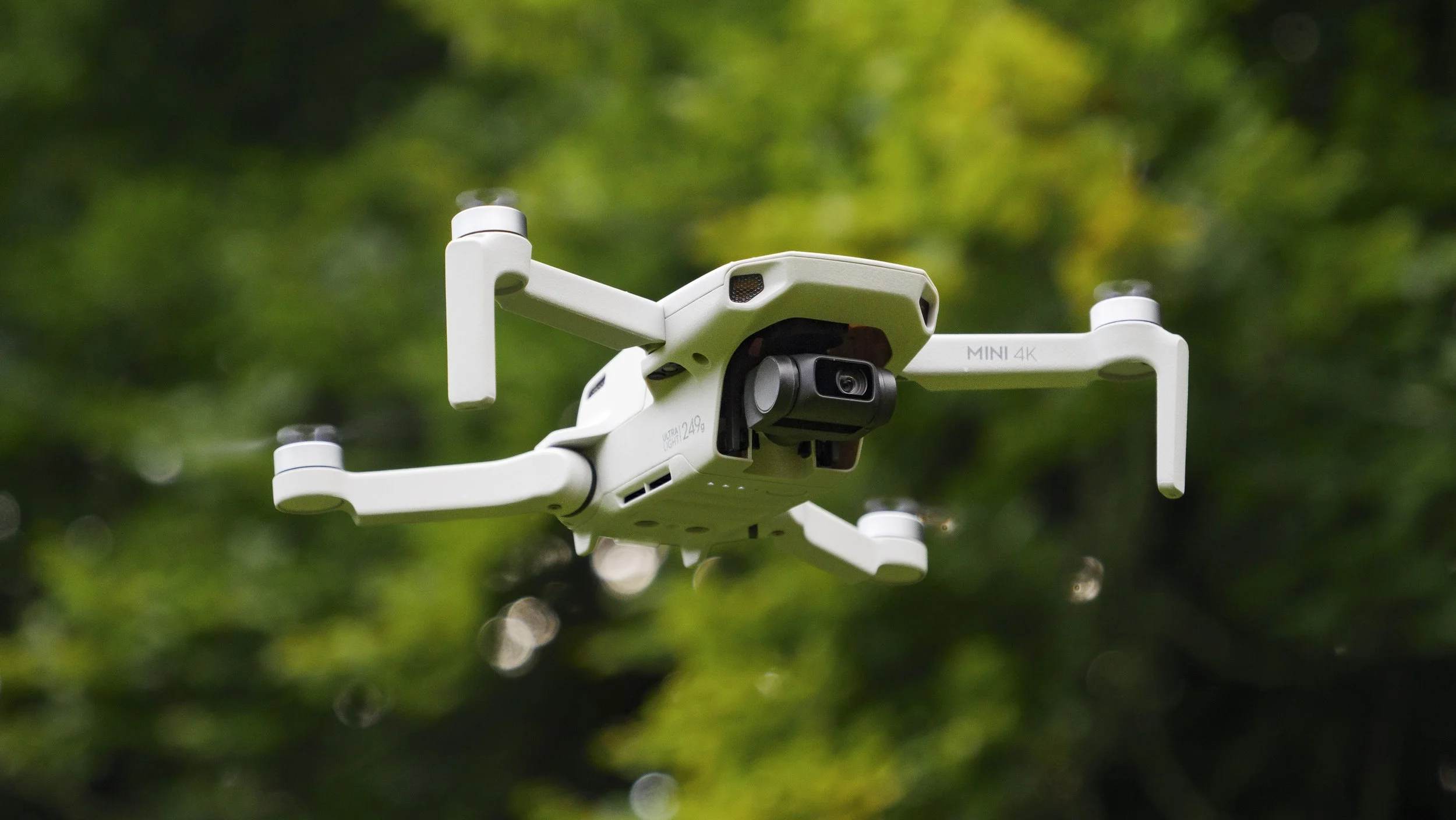 A white drone flying outdoors with a background of green foliage.