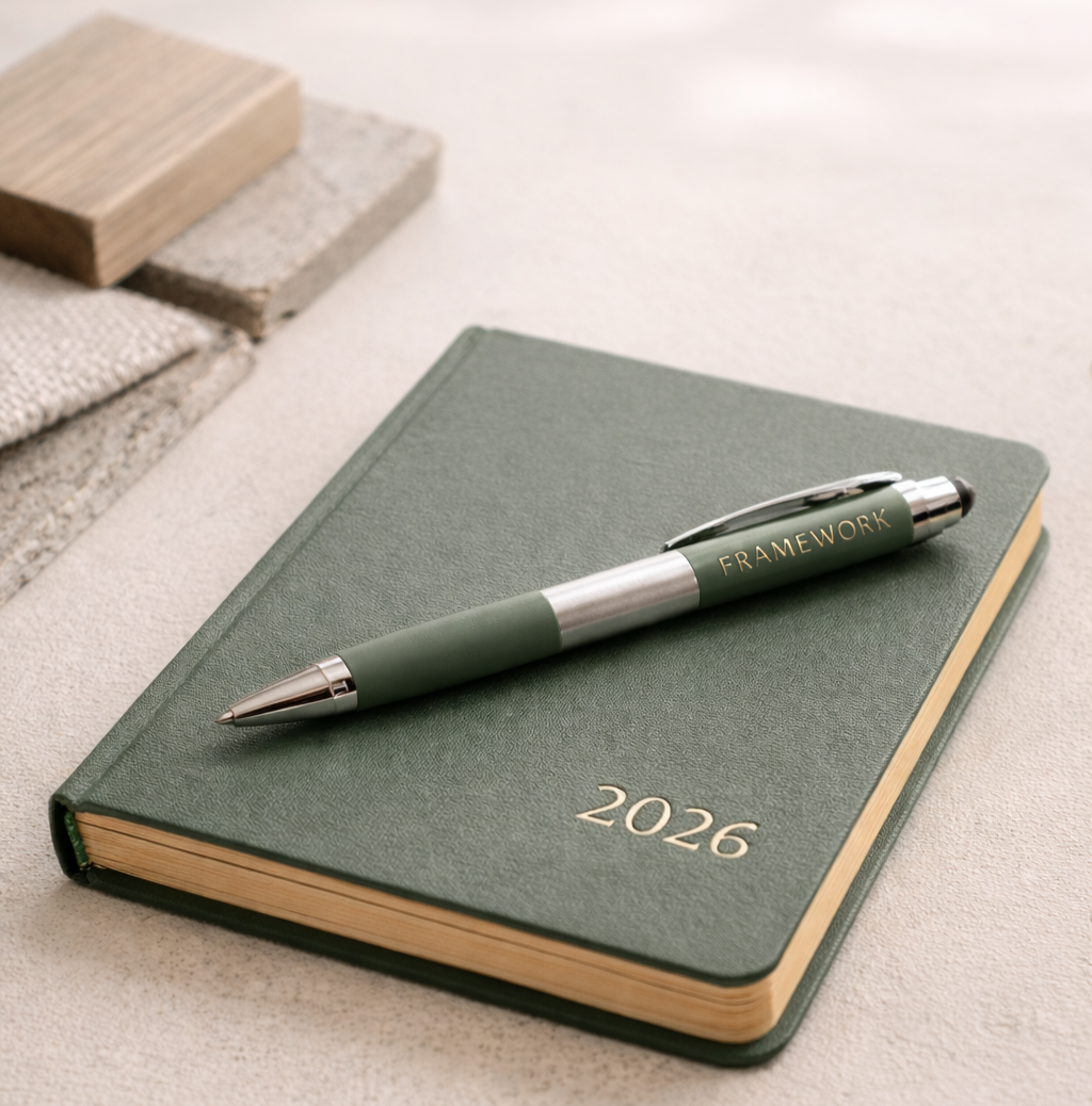 A green 2026 planner with a silver and green pen labeled "Framework" resting on top, placed on a light surface with fabric and wood sample swatches nearby.