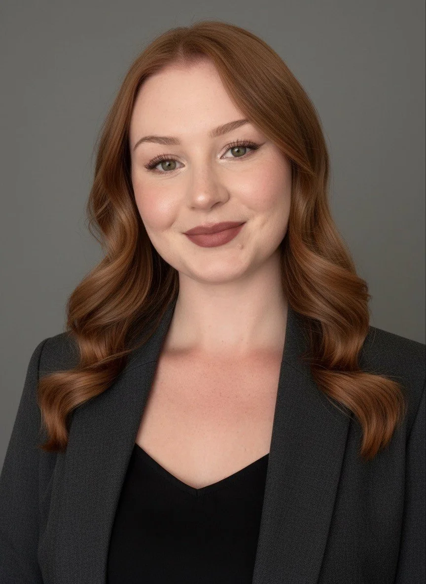 Portrait of a young woman with red hair, wearing a black top and a dark blazer, smiling softly against a gray background.