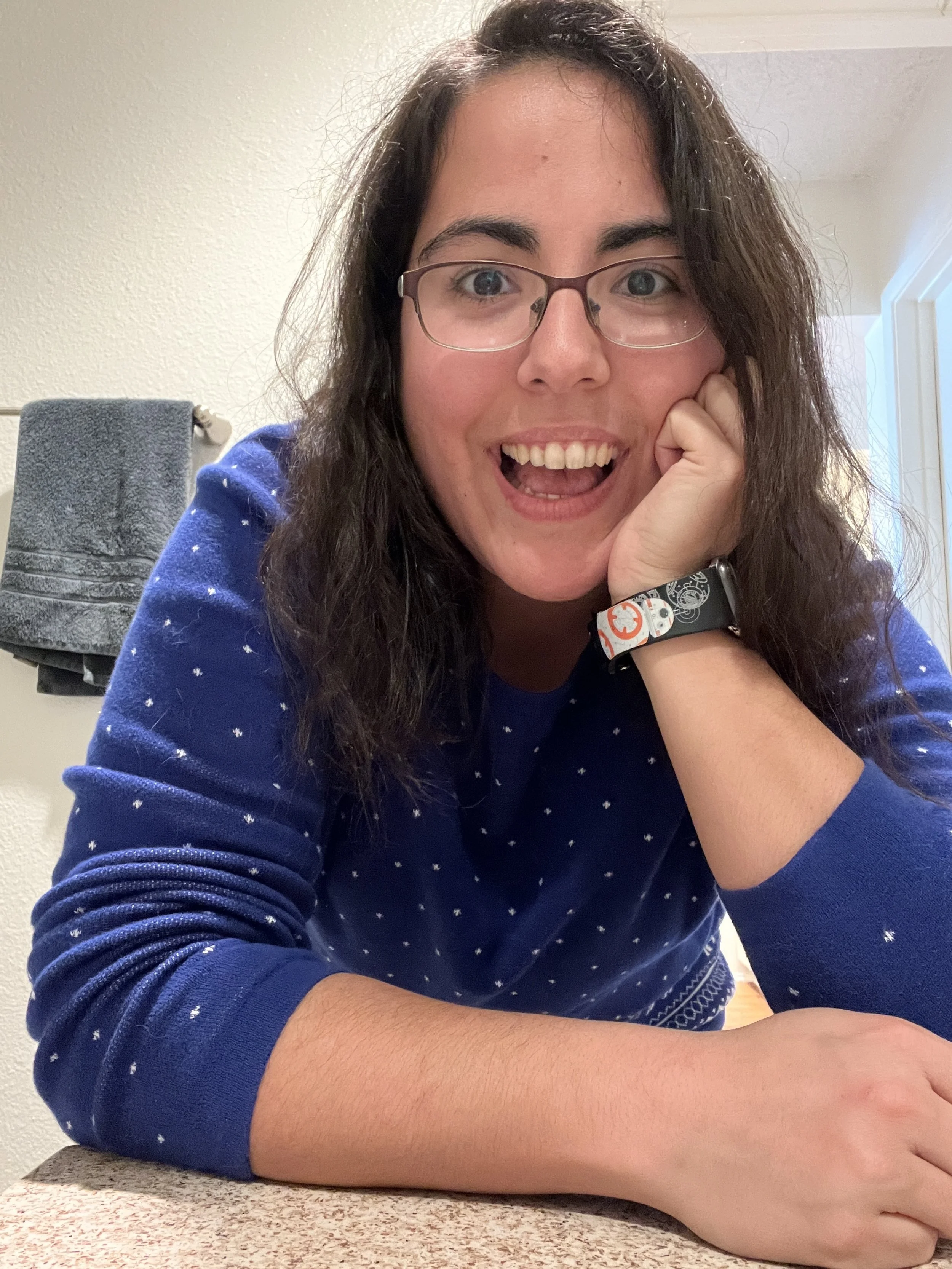 A woman with curly brown hair, glasses, and a blue sweater with white dots is smiling, resting her chin on her hand, in a room with towels in the background.