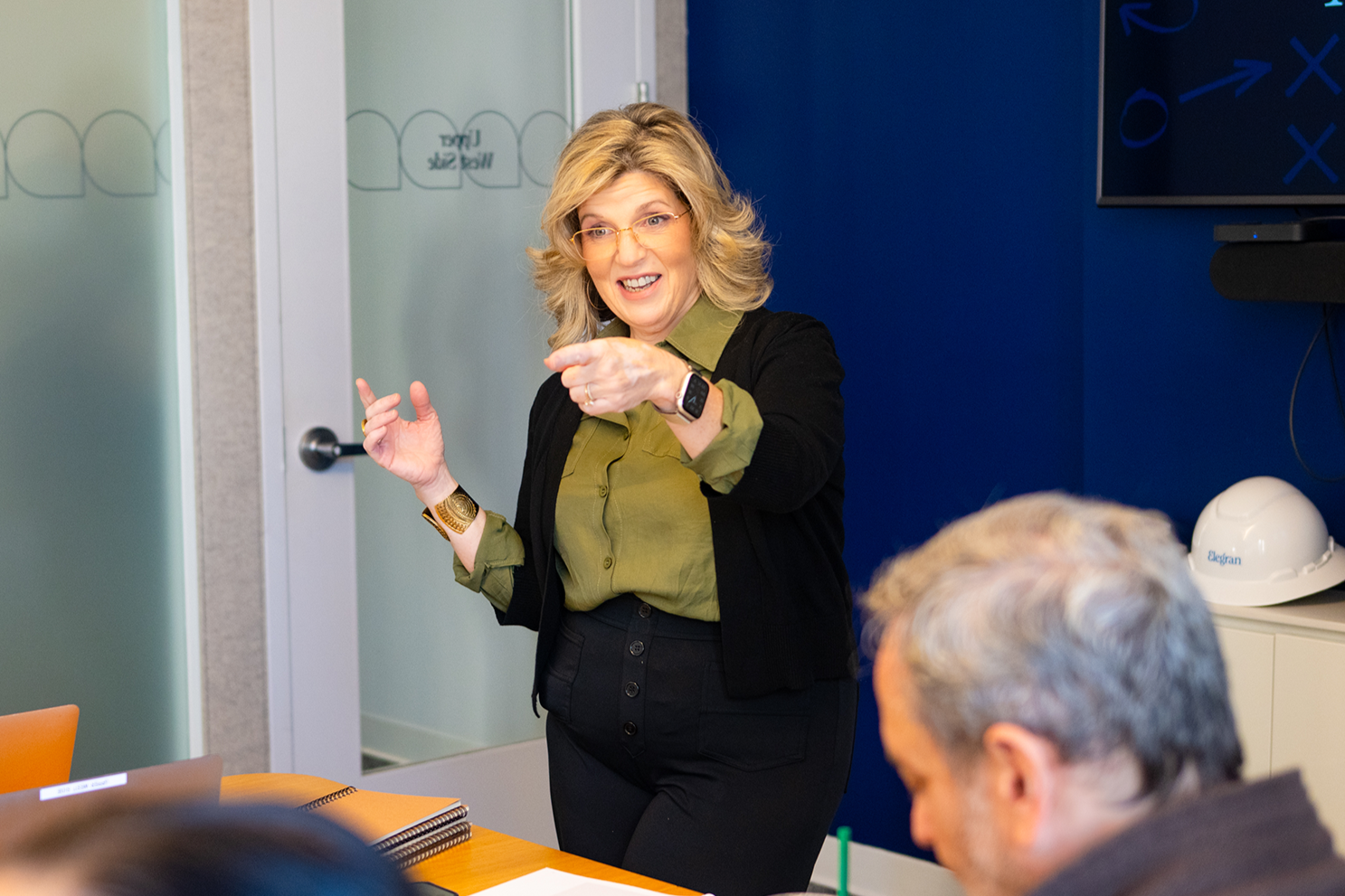 Amy is smiling while giving a presentation in a conference room teaching a real estate leadership  Masterclass. She is standing in front of a blue background showing diagrams and there is a rapt audience.