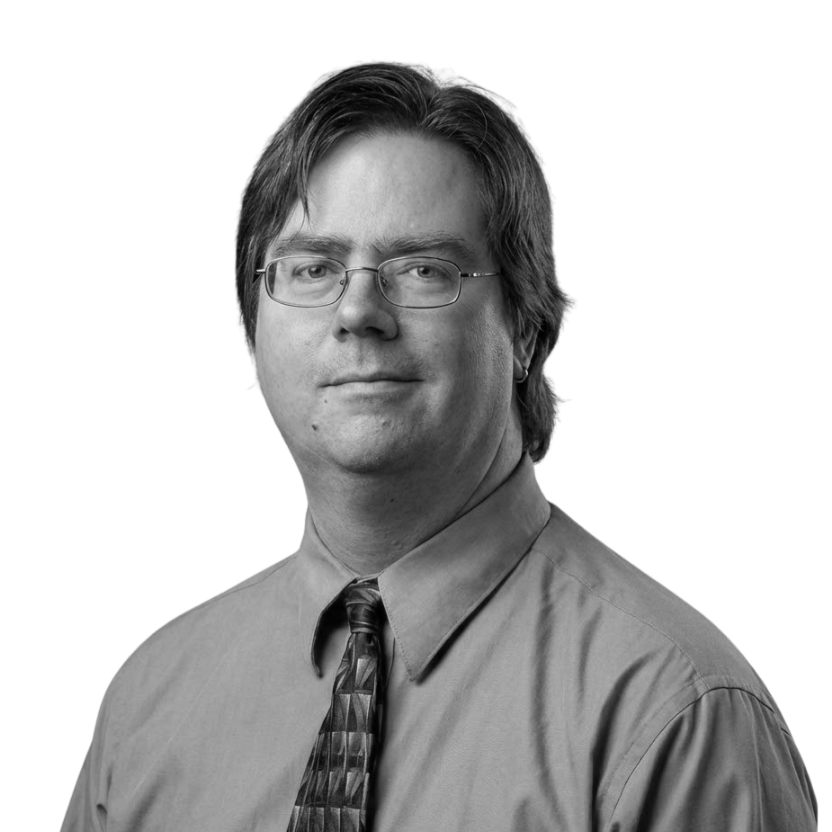 Black and white portrait of a man with glasses, medium-length hair, wearing a collared shirt and tie, looking directly at the camera.