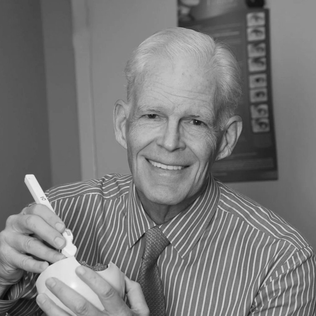 An elderly man with light hair wearing a striped shirt and tie, smiling while holding a spoon and a bowl, in a room with a bulletin board in the background.
