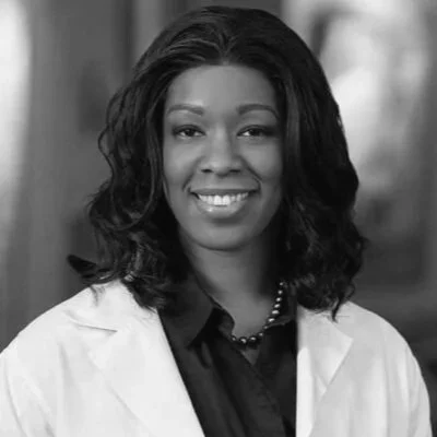 Portrait of a smiling African American woman with shoulder-length wavy hair, wearing a white lab coat over a dark shirt, in black and white.