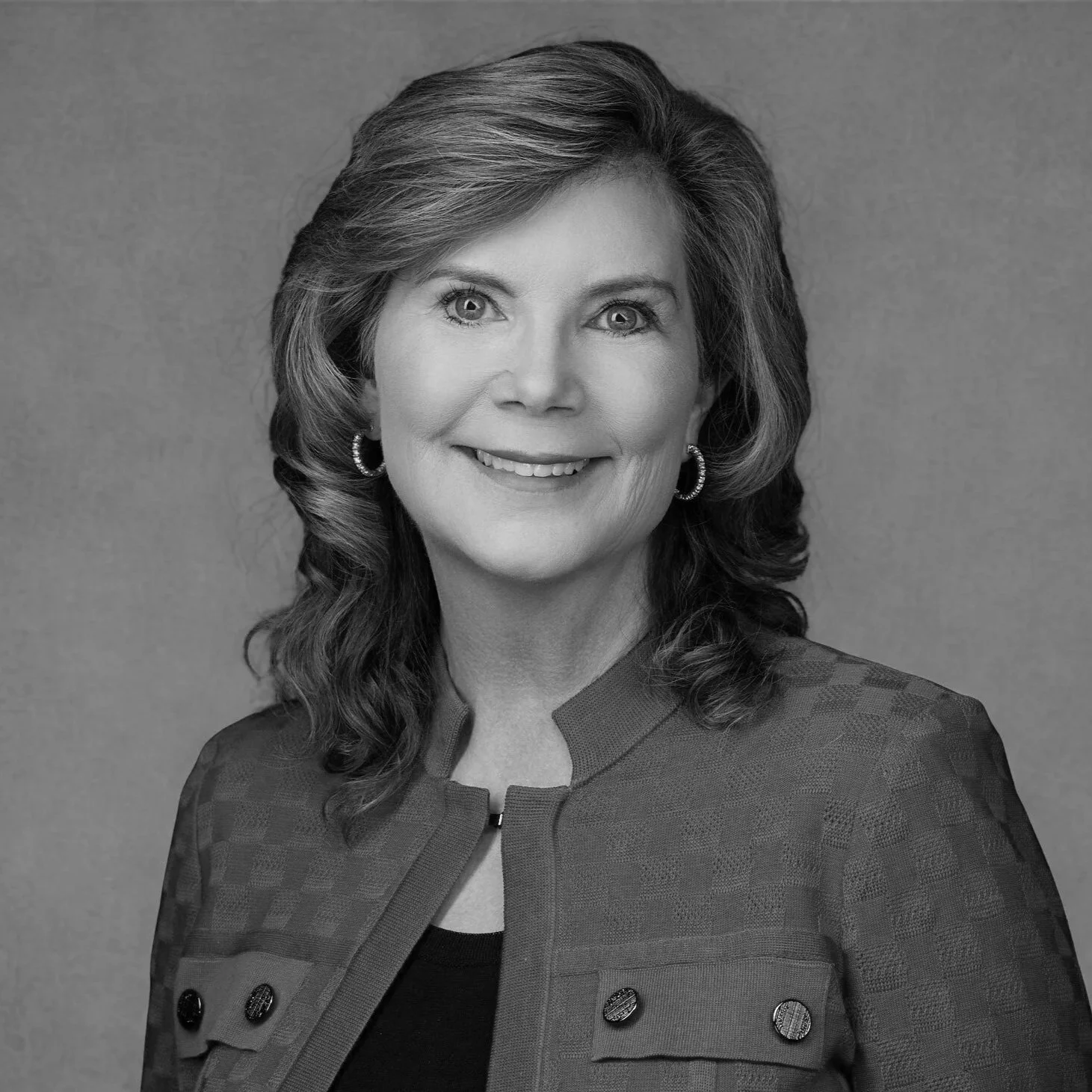 A black and white portrait of a middle-aged woman smiling, with wavy hair, hoop earrings, and a patterned jacket.
