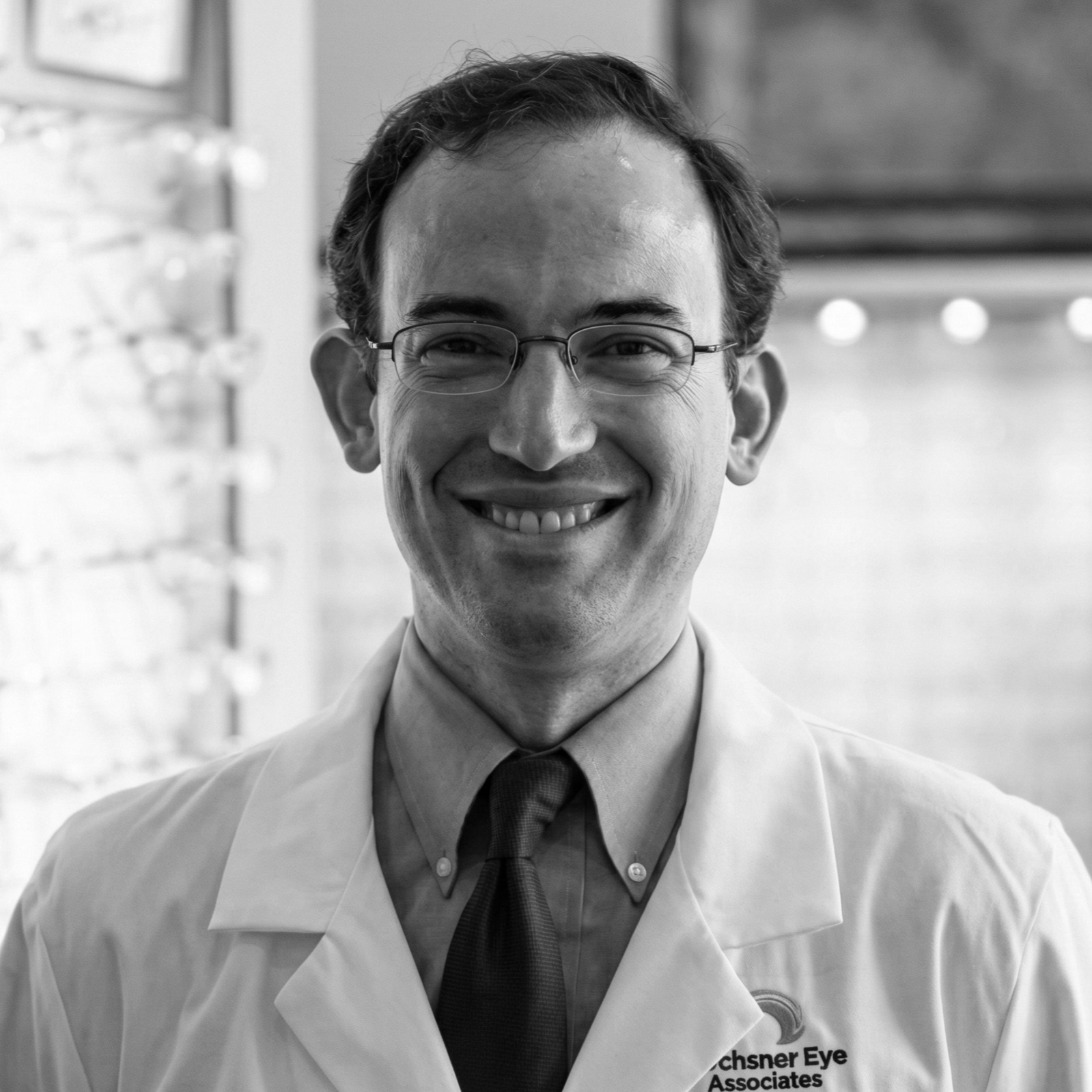 A smiling man in glasses wearing a lab coat with 'Ochsner Eye Associates' logo, standing indoors with a blurred background.