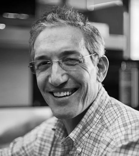 A smiling man wearing glasses and a checkered shirt, sitting indoors with blurred background.