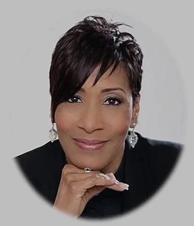 A woman with short dark hair, wearing earrings and a black outfit, smiling and resting her chin on her hand.