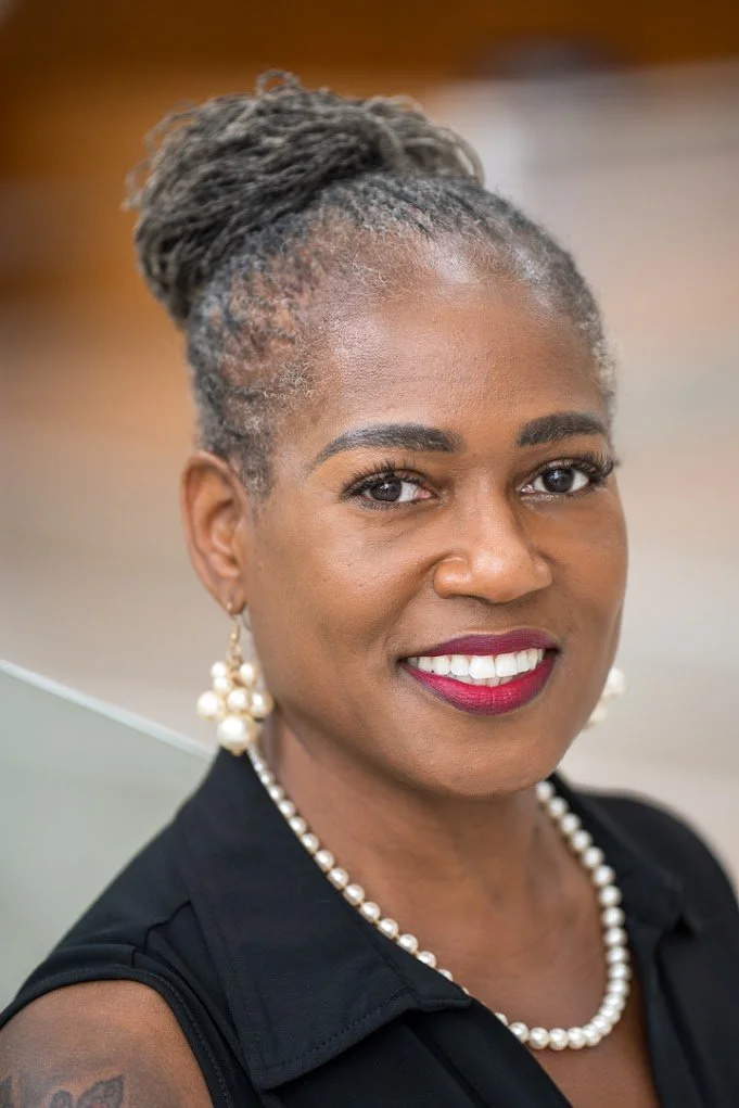 merican woman with dark, wavy hair, wearing pearl earrings, a pearl necklace, and a black blazer against a gray background.