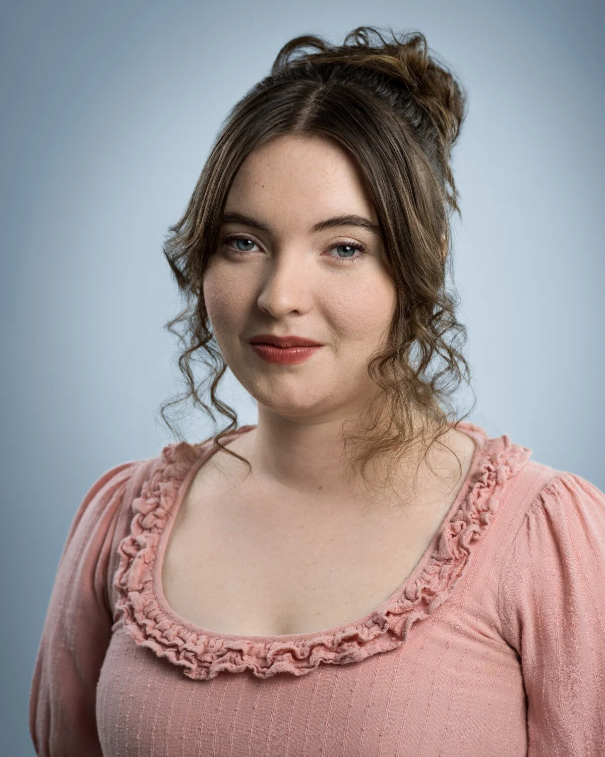 Skyela Jones in a pink period-style dress with a soft, knowing glimmer in her eyes. Her expression suggests a sharp, observational wit and a rich internal world, reflecting an essence of classical intelligence and modern-day grit.