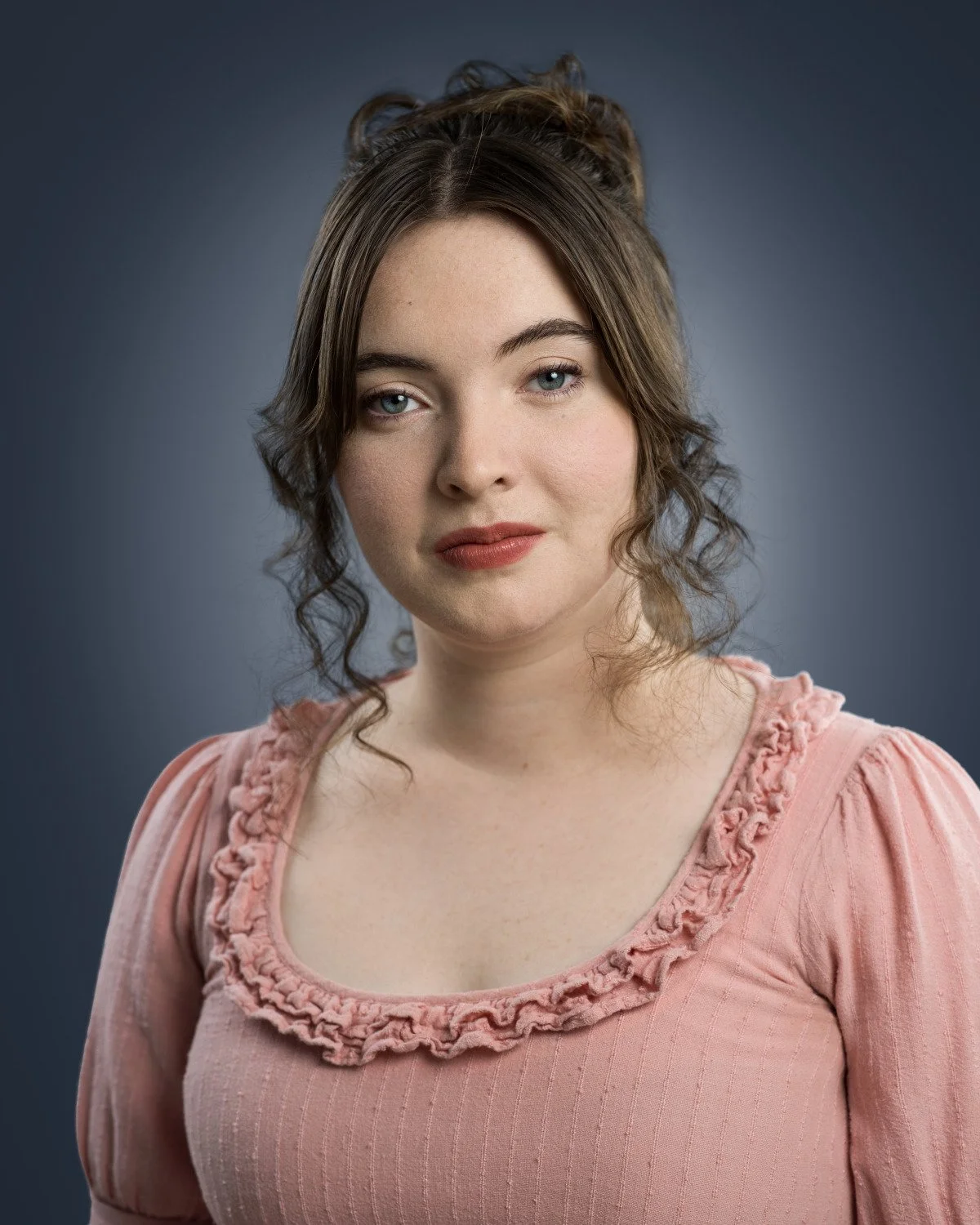 Theatrical headshot of Skyela Jones in a Regency-style pink dress. Her direct, intelligent gaze and subtle smile convey a sophisticated, grounded presence, perfectly suited to period-drama leads and classical Shakespearean roles.
