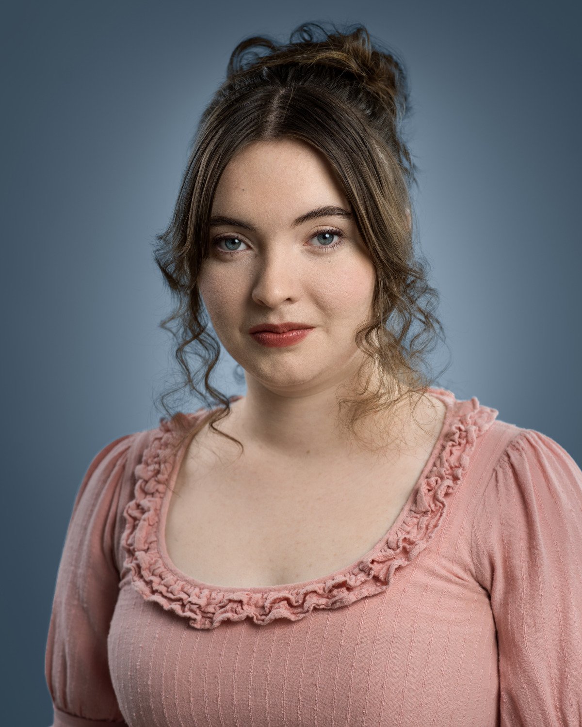 Theatrical headshot of Skyela Jones with a steady and enigmatic gaze. Styled in a pink ruffled gown with an elegant updo, she portrays a deeply observant, authoritative character in high-stakes period dramas.
