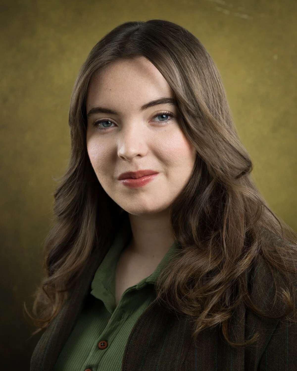 Warm, approachable headshot of Skyela Jones with blue eyes, wavy hair, wearing a green polo and textured blazer. She has a soft smile and a friendly gaze, suited for commercial or professional "everyman" roles: med student, caretaker, best friend.