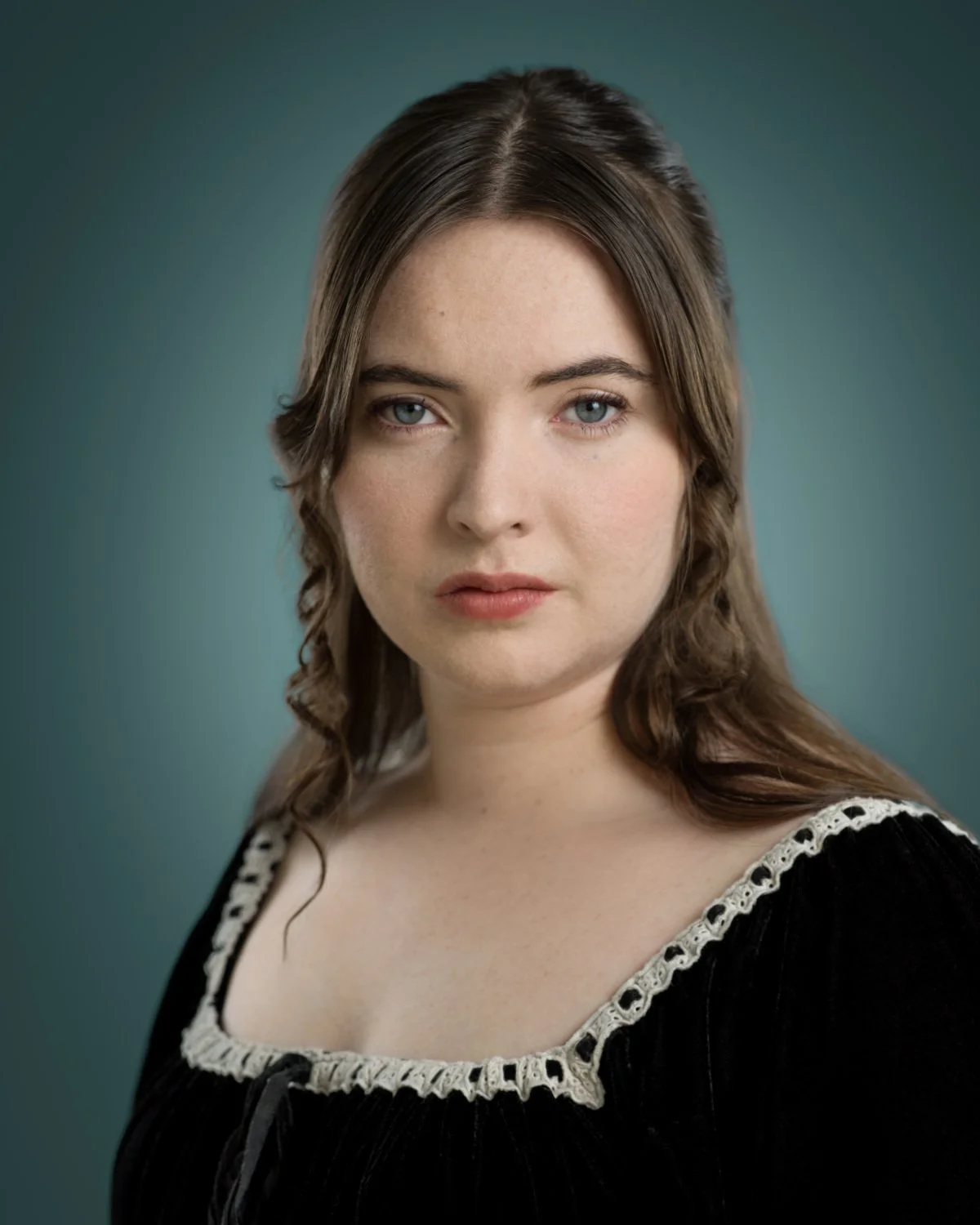 Dramatic period headshot of Skyela Jones wearing a black velvet dress with white lace trim. Her brown hair is styled into classic curls, and her expression is solemn and intense, reflecting a Shakespearean or Gothic-drama aesthetic.
