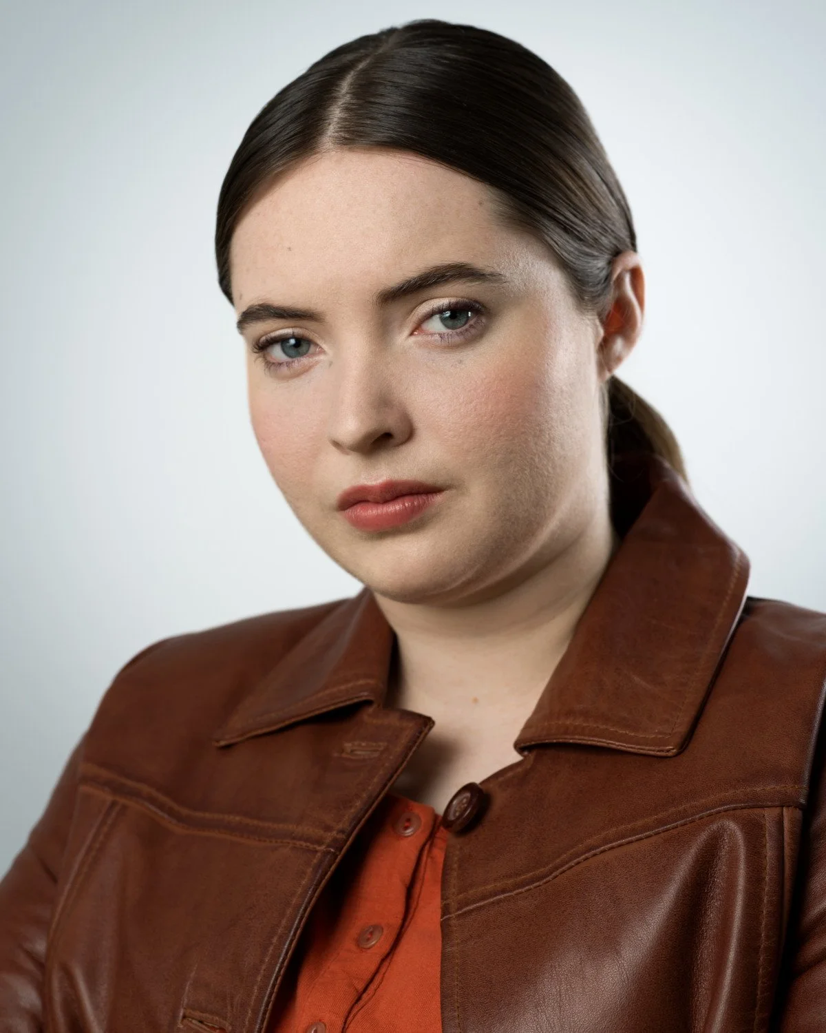 Professional headshot of Skyela Jones with blue eyes, brown hair pulled back, wearing a brown leather jacket over an orange top. She has a serious, grounded expression, suitable for modern procedural or dramatic television roles.