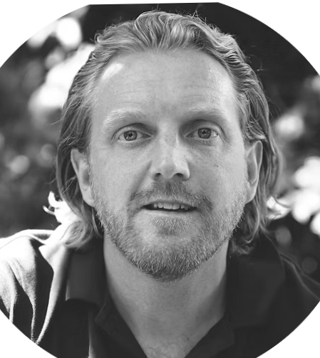 A black-and-white portrait of a man with wavy hair and a beard, smiling and looking at the camera.