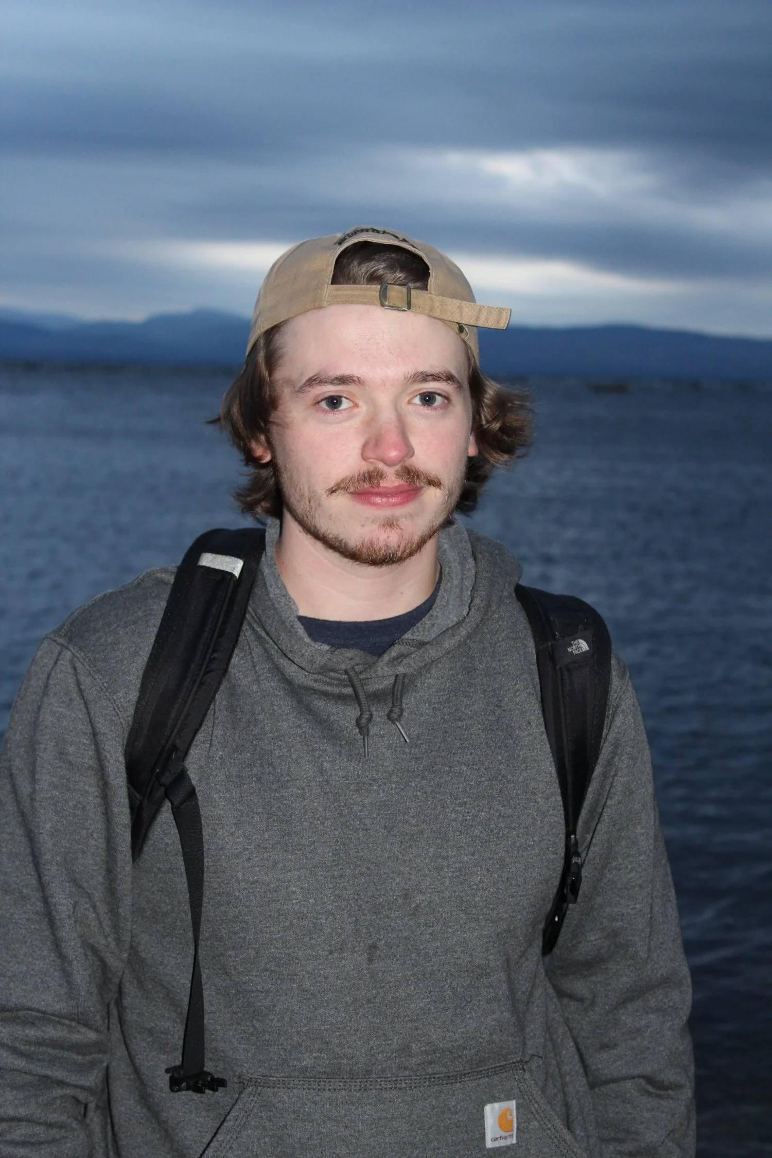 A young man with light brown hair, a mustache, and a beard stands outdoors by a body of water with mountains in the background, wearing a gray hoodie, a beige cap backwards, and a black backpack.
