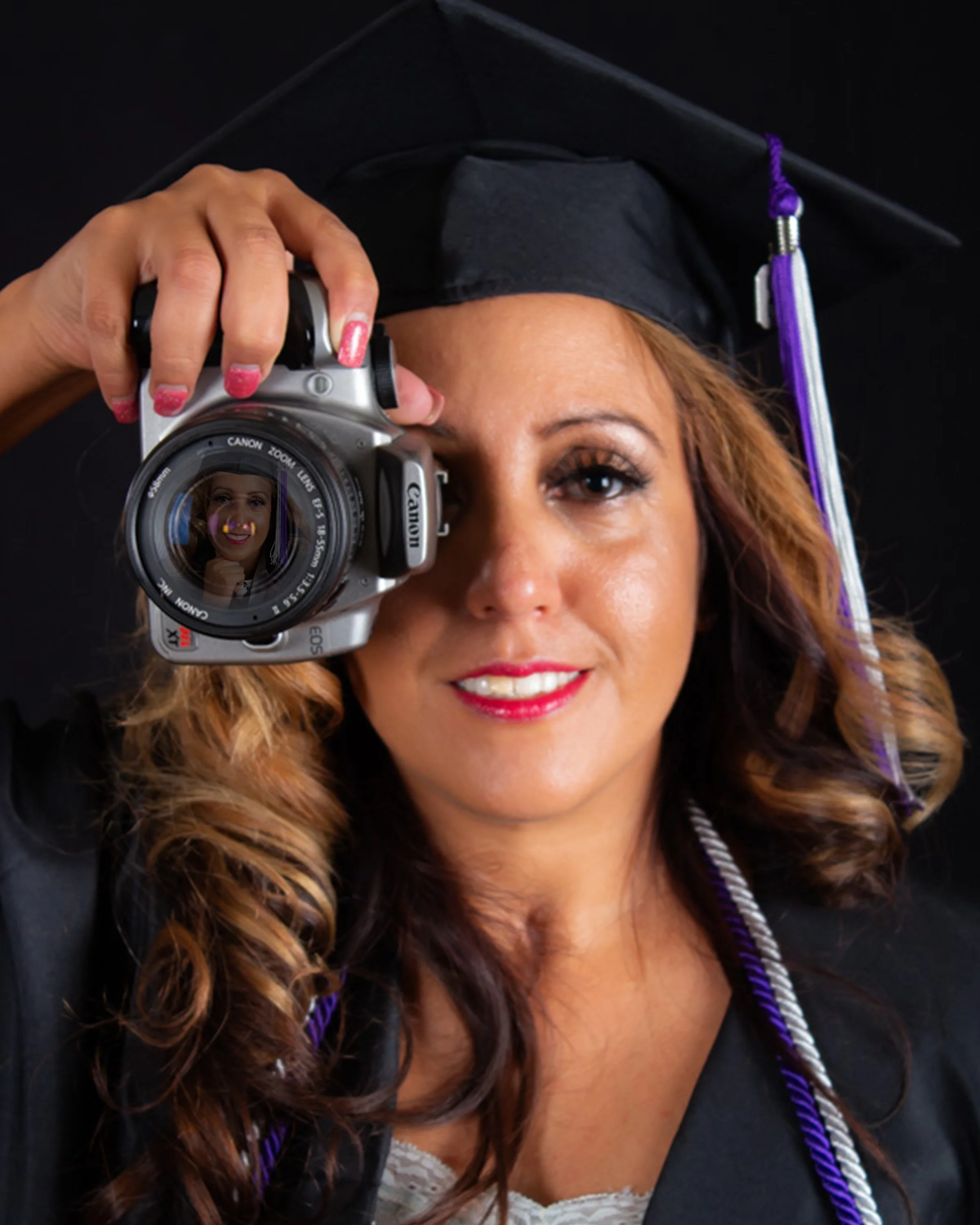 Woman in graduation cap and gown taking a photo with a camera, reflection of her face visible in the camera lens.